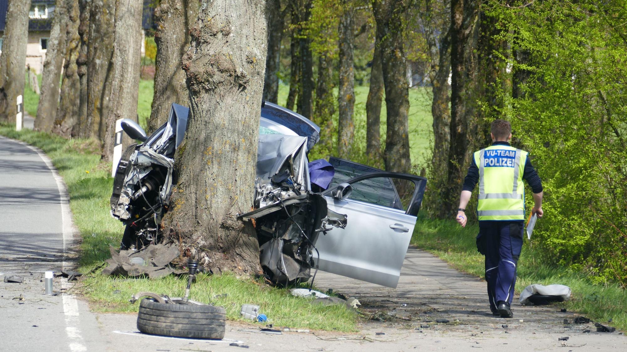 Rechts ein Polizist von hinten, in der Mitte ein Baumstamm, gegen den ein Pkw gefahren ist, herumliegende Trümmerteile