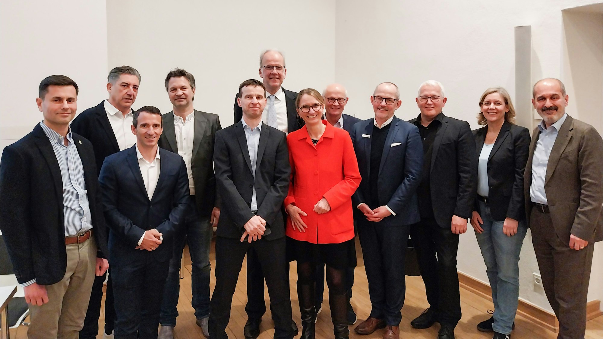 Zehn Männer in dunklen Jackets und zwei Frauen, eine in rotem Mantel, lächeln in die Kamera