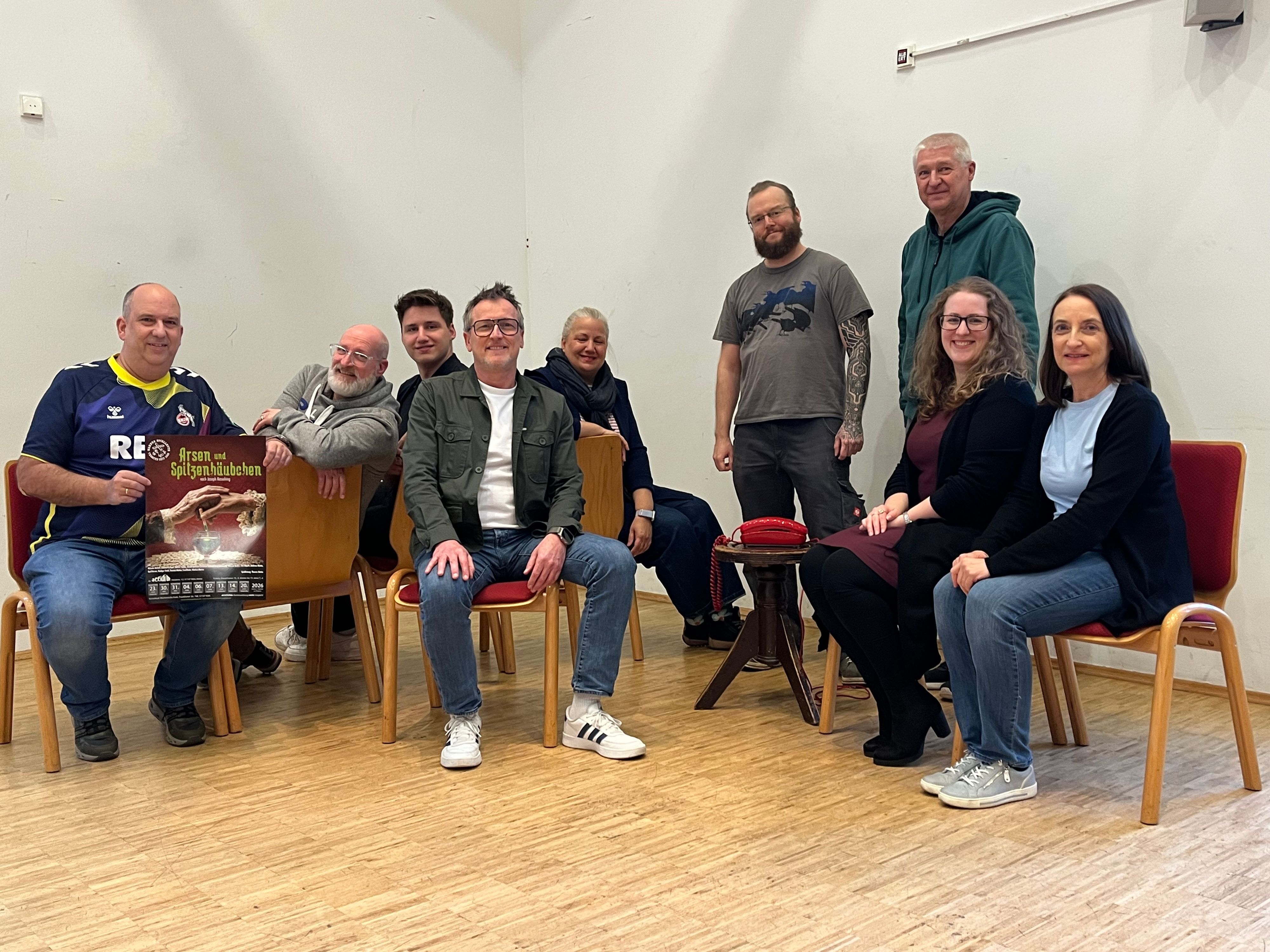 Neun Menschen sitzen auf Stühlen oder stehen in einem Pfarrzentrum.