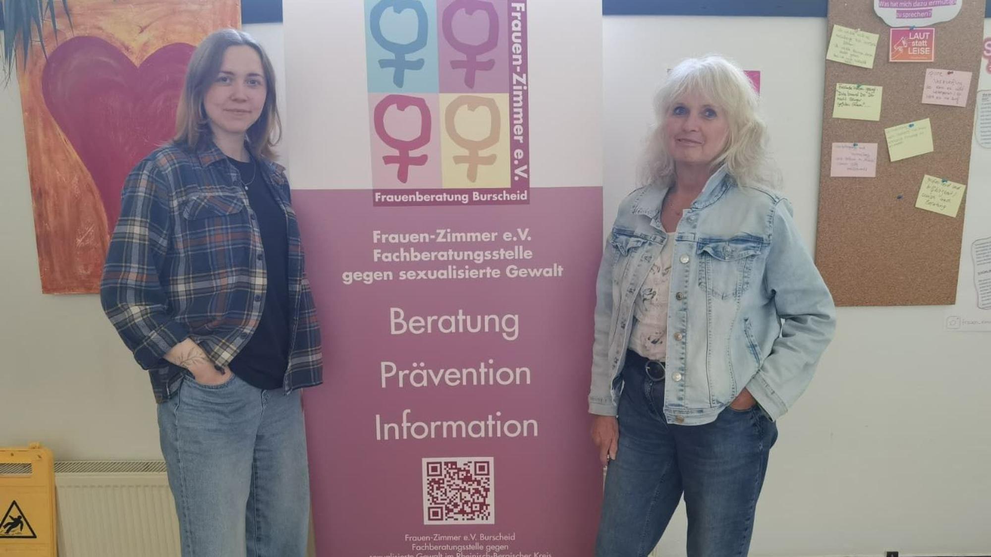 Zwei Frauen stehen links und rechts von einem Banner, das Kontaktmöglichkeiten zu einer Beratungsstelle gegen sexualisierte Gewalt aufzeigt.