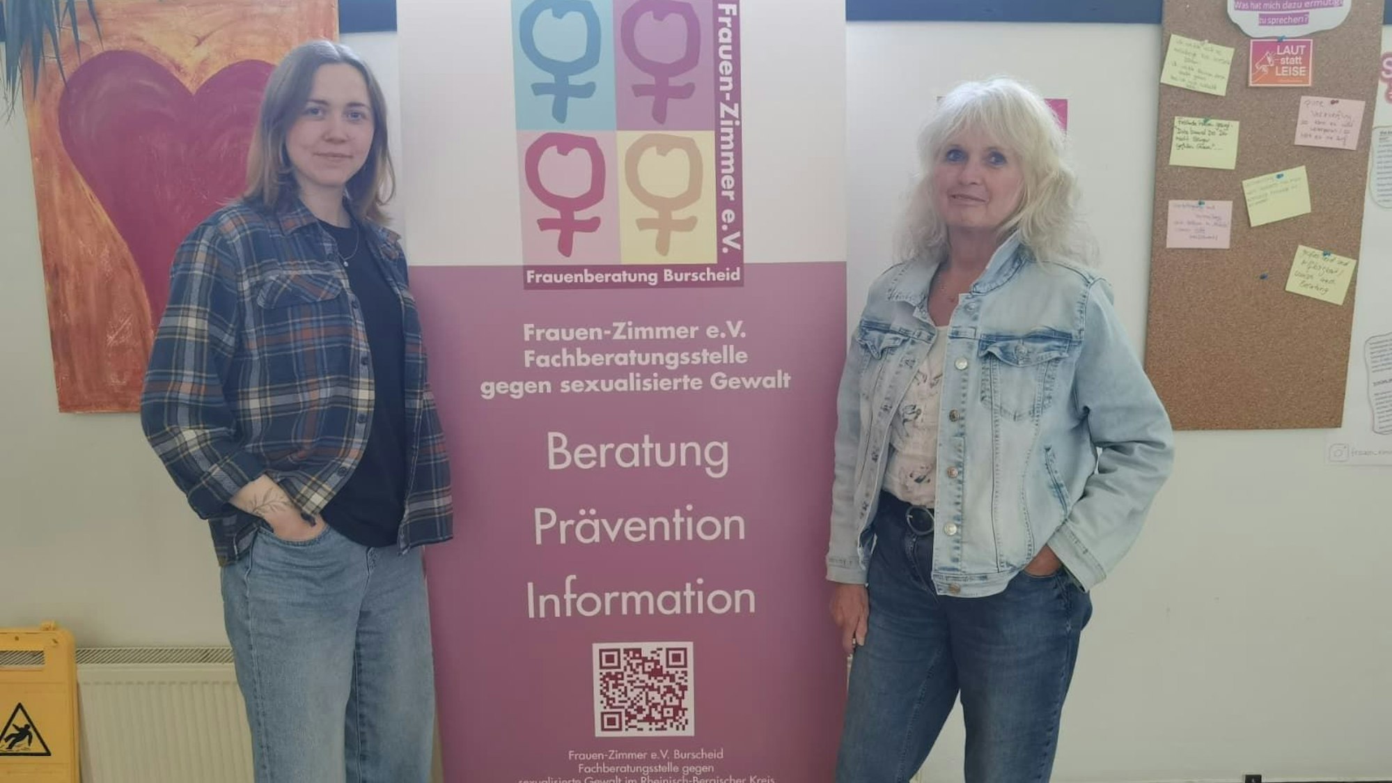 Zwei Frauen stehen links und rechts von einem Banner, das Kontaktmöglichkeiten zu einer Beratungsstelle gegen sexualisierte Gewalt aufzeigt.