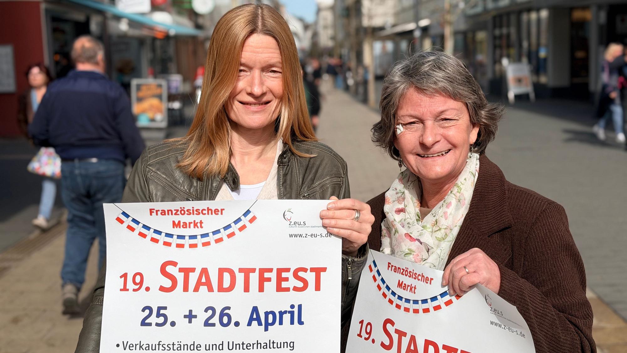 Die beiden Frauen stehen in der belebten Fußgängerzone, in den Händen halten sie jeweils ein Plakat für das Stadtfest.