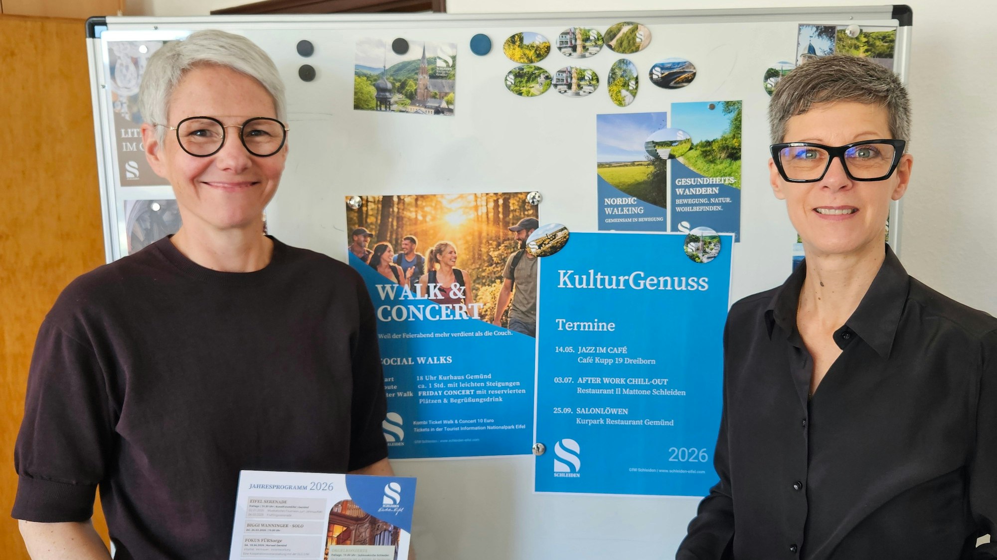 Zwei Frauen stehen vor einer Tafel mit Werbeplakaten.