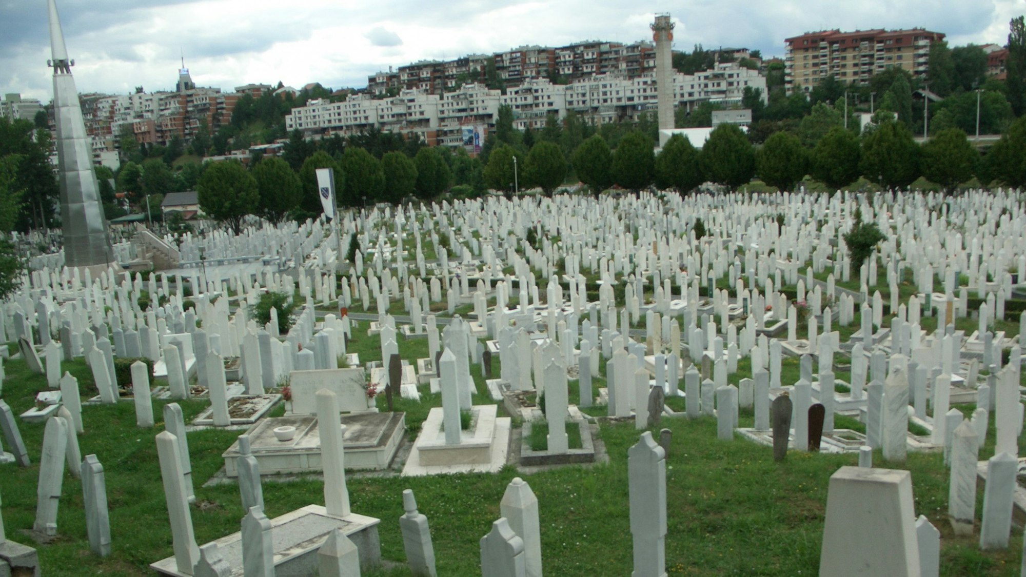 Ein Friedhof mit vielen weißen Kreuzen auf einer Wiese. Dahinter eine Säule mit olympischen Ringen.