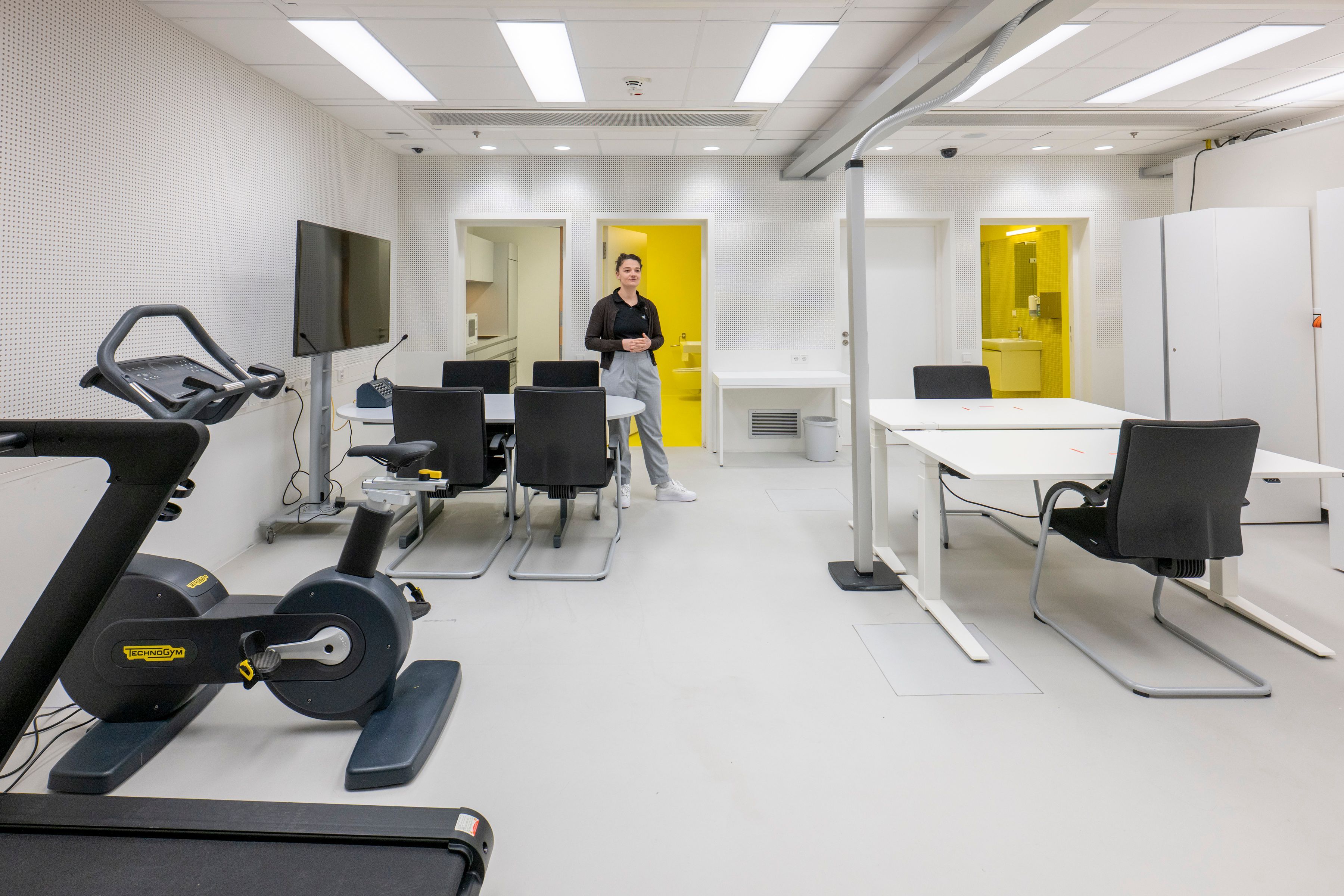 Der Aufenthaltsraum der SOLIS-Studie: Es gibt ein Laufband, ein Fahrradergometer, zwei Tische und einen Fernseher. Im Hintergrund sieht man den Eingang zu zwei Bädern und der Küche.