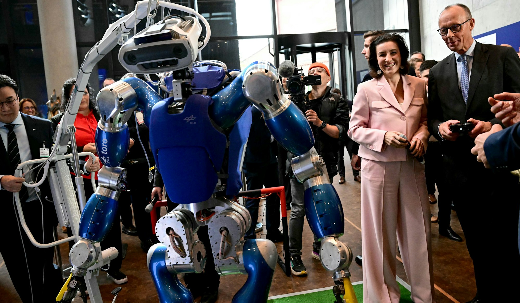 Das Foto zeigt Bundesforschungsministerin Dorothee Bär (Zweite von rechts= und Bundeskanzler Friedrich Merz (rechts) mit dem humanoiden Laufroboter Toro des Deutschen Zentrums für Luft- und Raumfahrt.