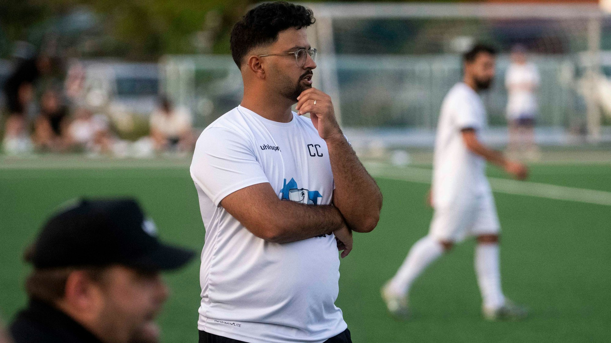Bessenichs Trainer Can Celik steht bei einem Spiel im weißen T-Shirt an der Außenlinie.