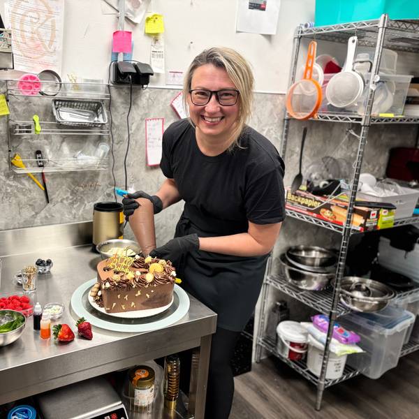 Eine Frau mit blonden Harren steht in schwarz gekleidet in einer Küche und verziert eine Schokoladen-Torte.