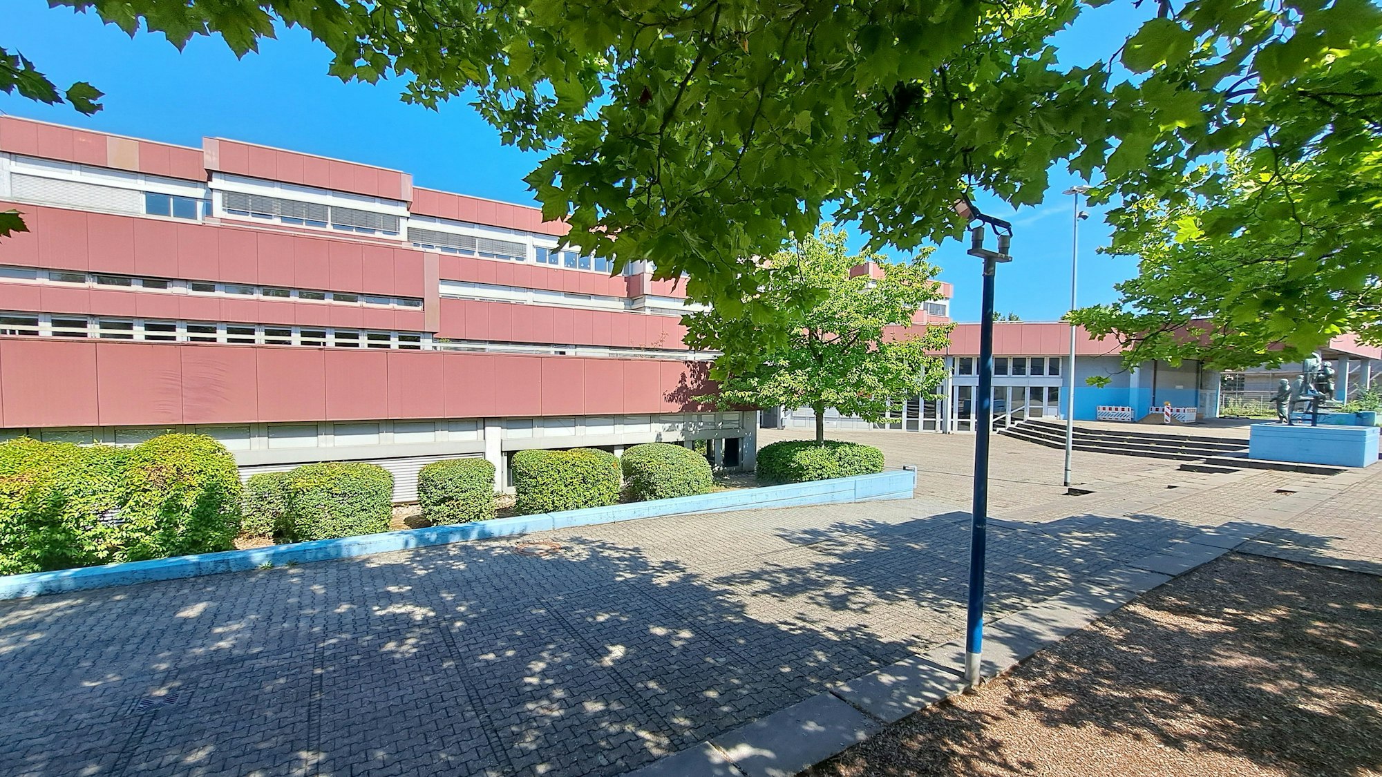Die Aufnahme zeigt das Kopernikus-Gymnasium im Niederkasseler Schulzentrum Nord. Im Vordergrund werfen Bäume einen Schatten auf Pflastersteine.