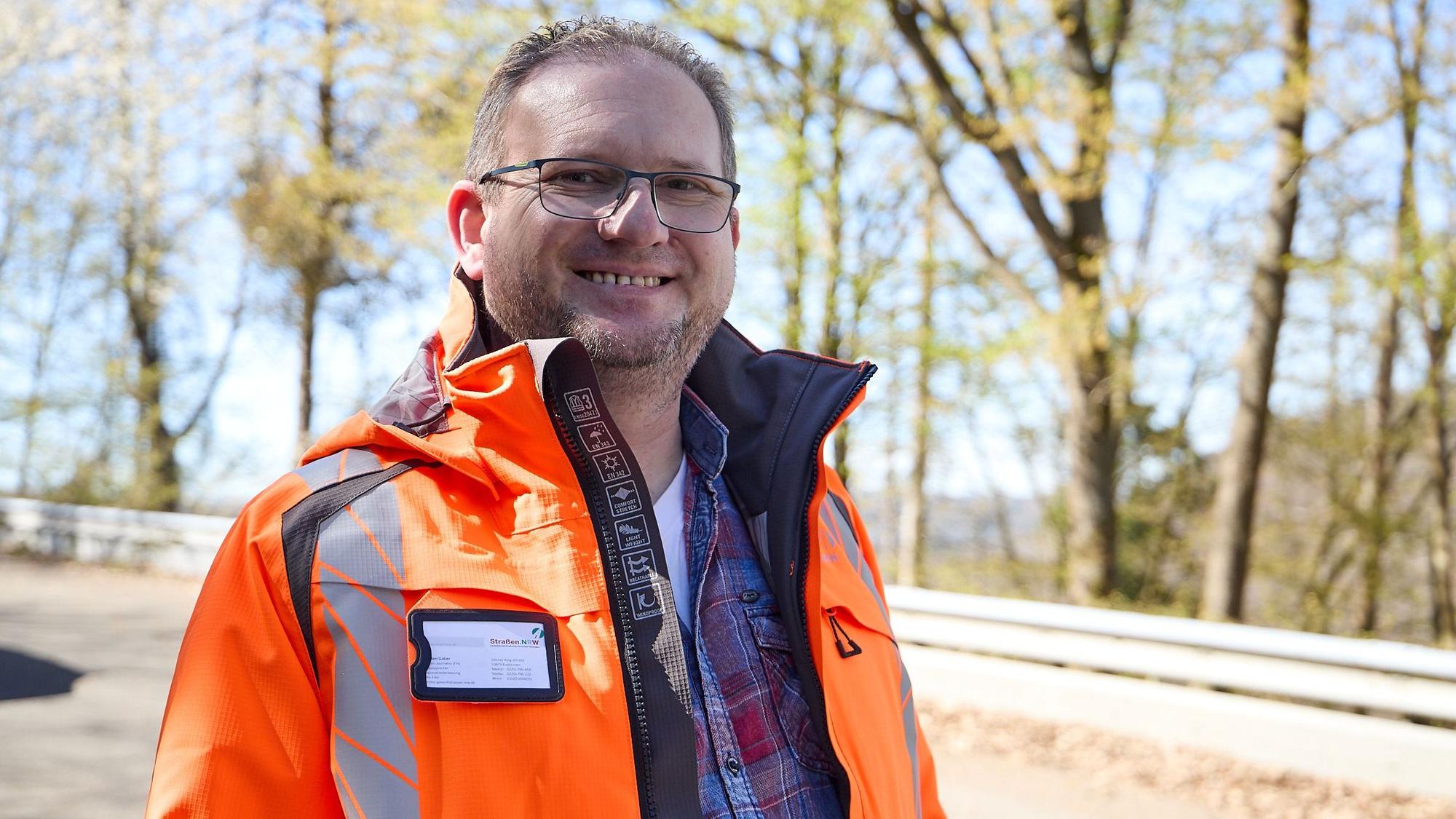 Torsten Gaber, Sprecher vom Landesbetrieb Straßen NRW, trägt eine orangefarbene Jacke. Er steht auf einer Straße, im Hintergrund sind eine Leitplanke und Bäume zu sehen.