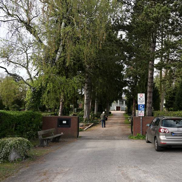 Der Eingang eines Friedhofes mit hohen Bäumen. Auf dem Weg geht ein älterer Mann.