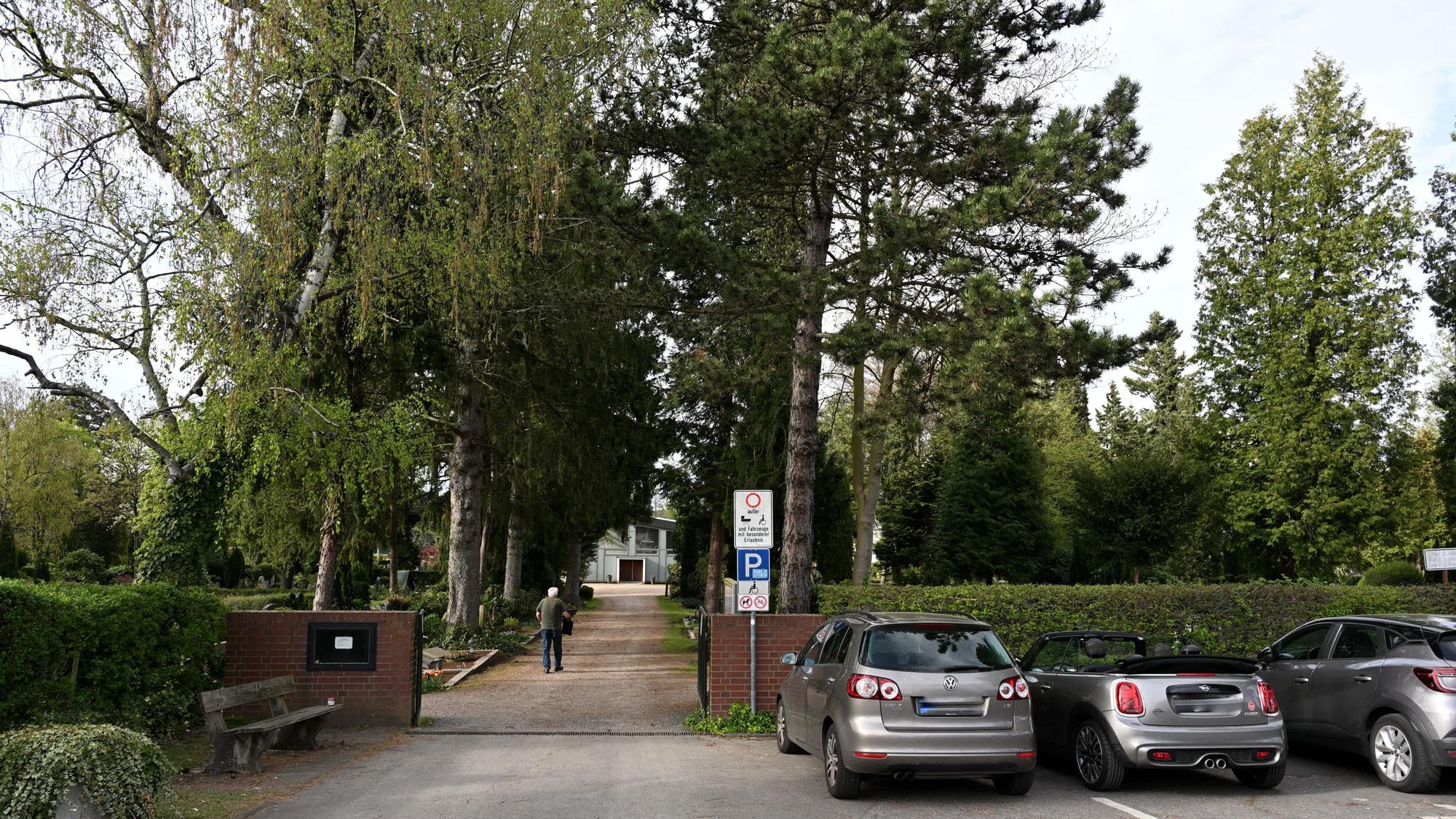 Der Eingang eines Friedhofes mit hohen Bäumen. Auf dem Weg geht ein älterer Mann.