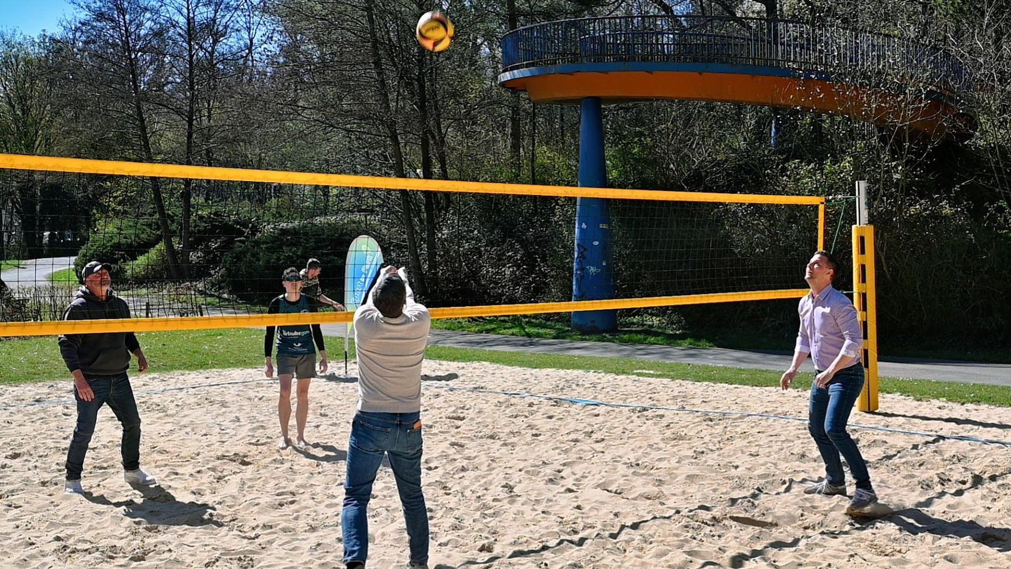 Auf einem Spielfeld mit Sandboden sieht man vier Menschen Beachvolleyball spielen.