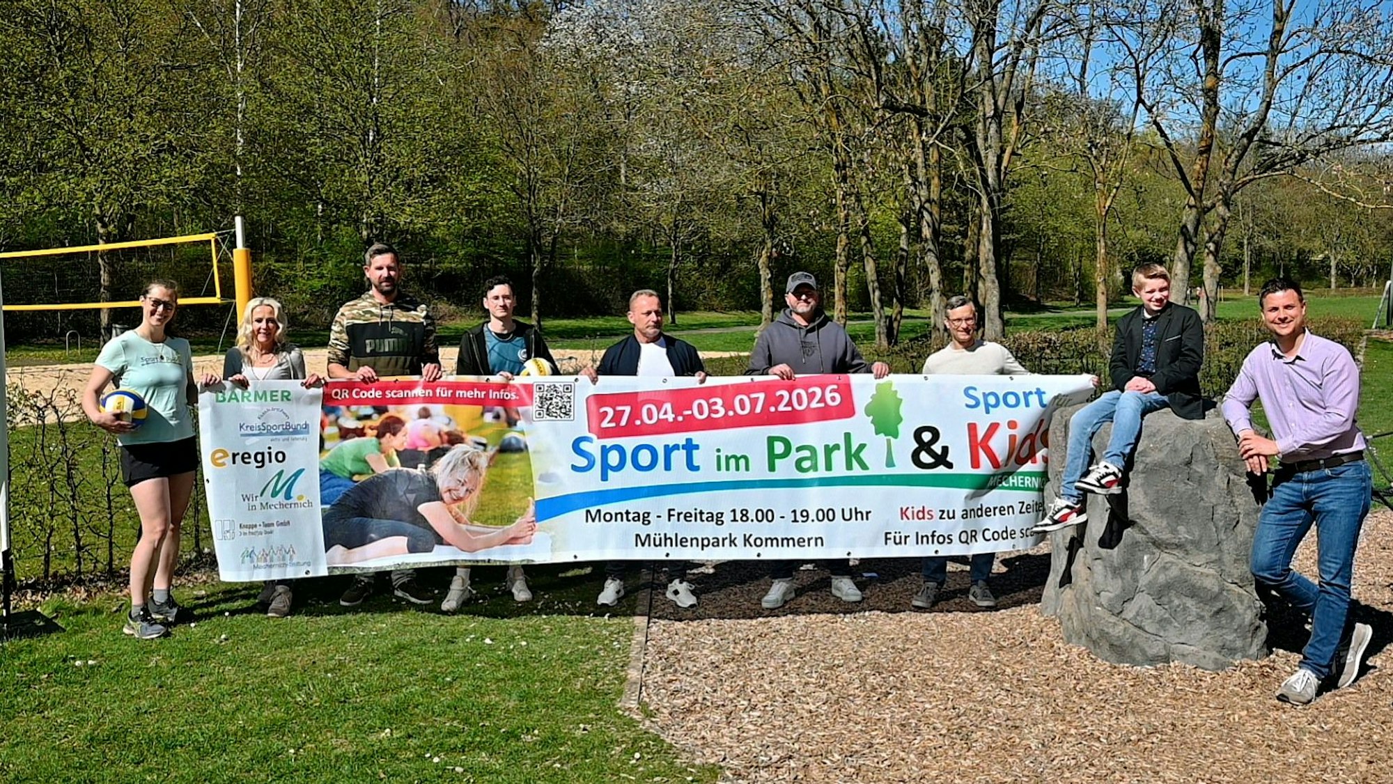 Im Mechernicher Mühlenpark stehen Mitglieder des Organisations- und Trainerteams hinter einem Banner, der das „Sport im Park“-Projekt bewirbt.