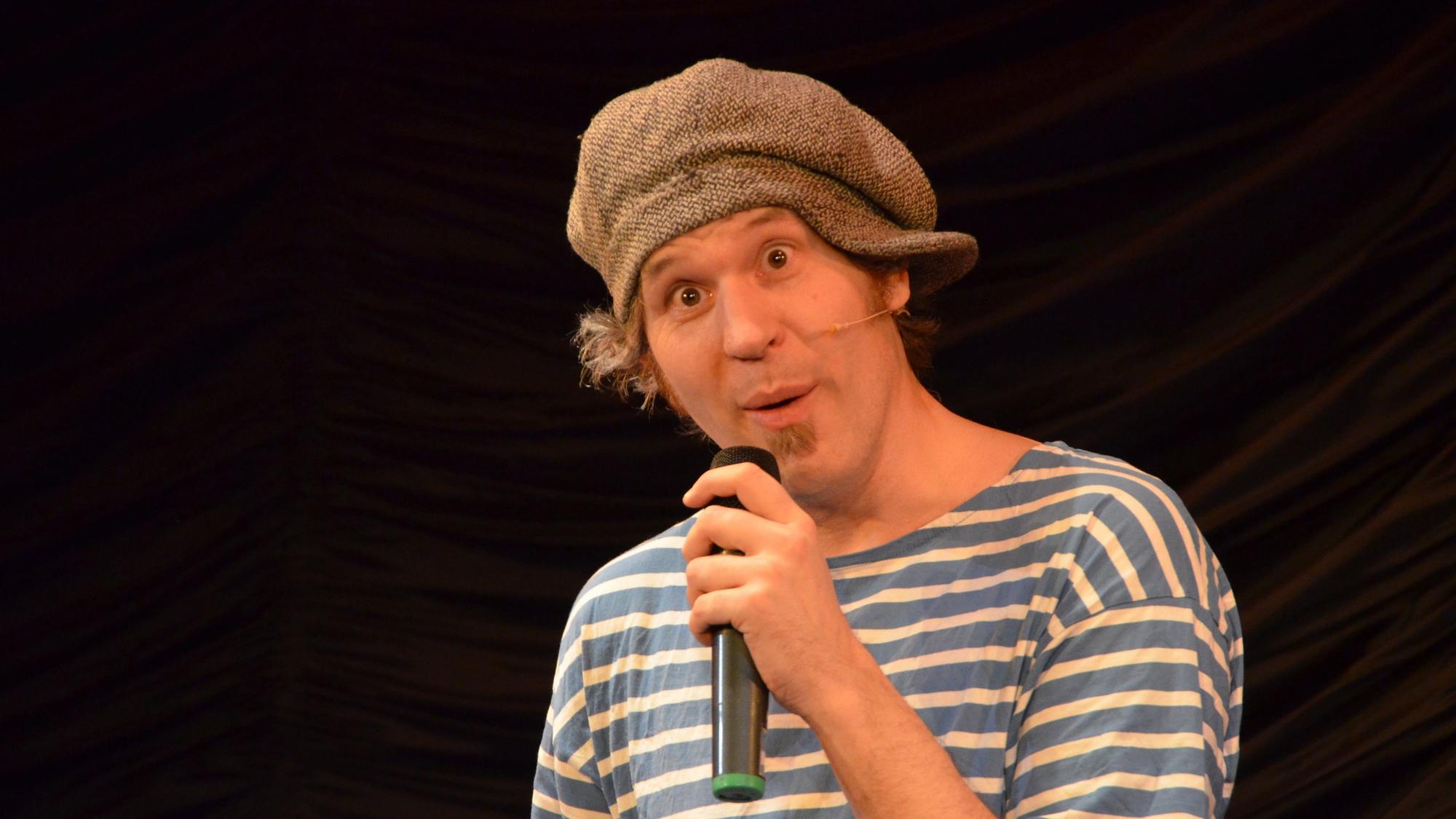 Drop Bert hat ein Streifen-Shirt an und eine Kappe schief auf dem Kopf. Er spricht in sein Mikro, das er in der Hand hält und reißt die Augen weit auf.