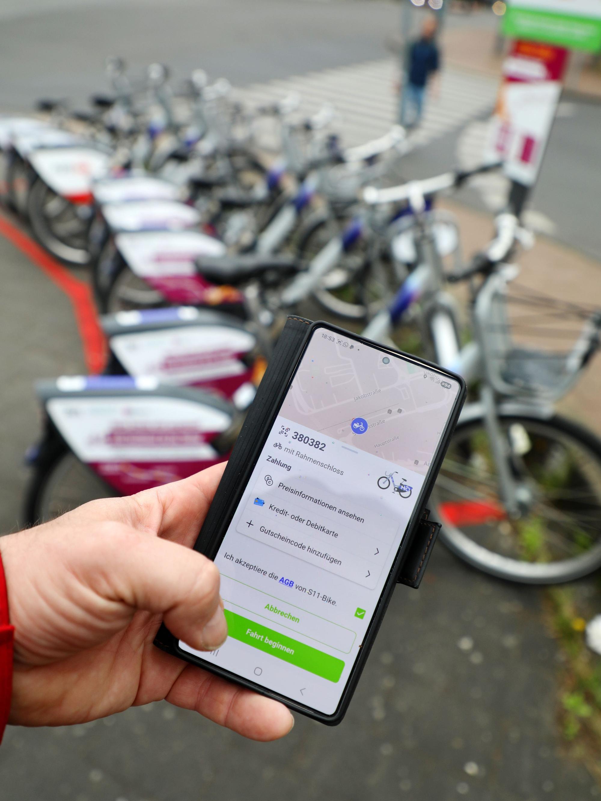 Eine Hand hält ein Handy mit einer App, mittels derer eines von mehreren Fahrrädern im Hintergrund ausgeliehen wird.