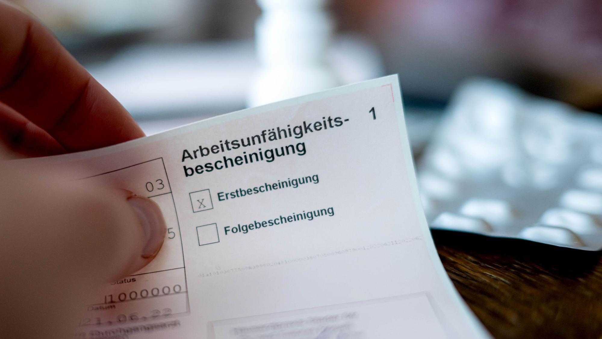 Eine Person hält eine ausgefüllte Arbeitsunfähigkeitsbescheinigung in der Hand. Im Hintergrund ist unscharf ein Tisch mit Medikamenten zu sehen. (Symbolbild)