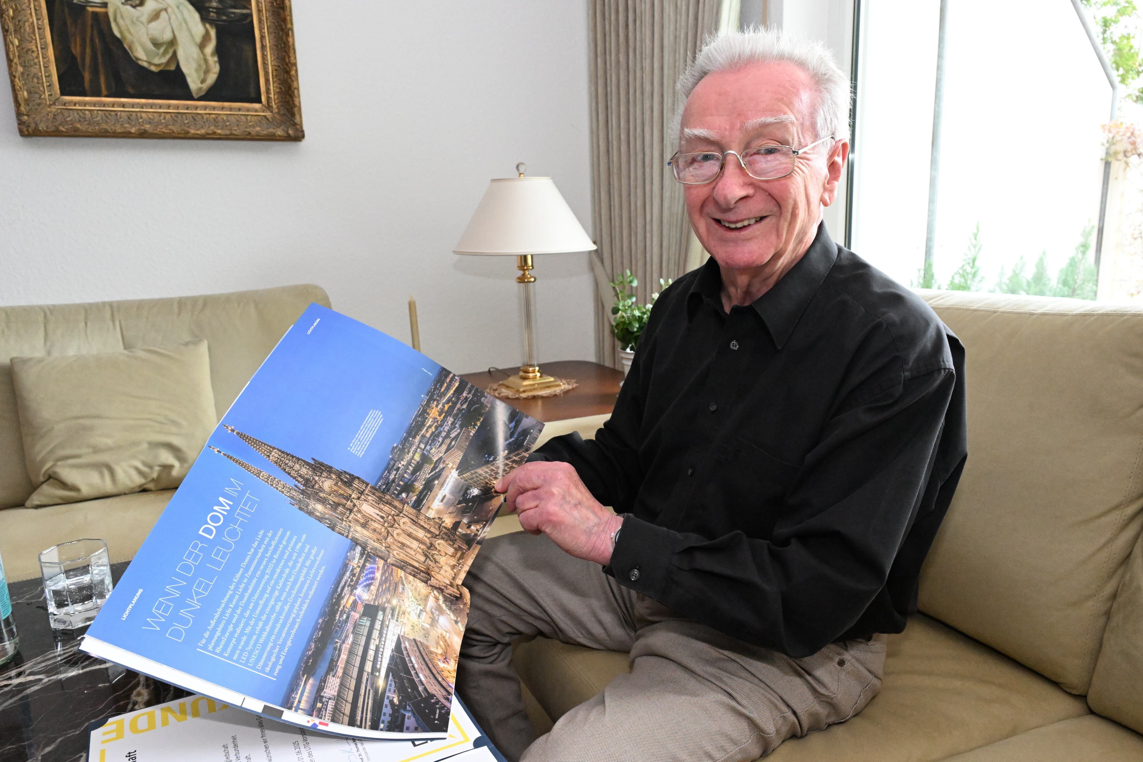 Ein älterer Herr sitzt auf einem Sofa und hält ein Heft mit einem Hochglanzfoto vom beleuchteten Klölner Dom in die Kamera.