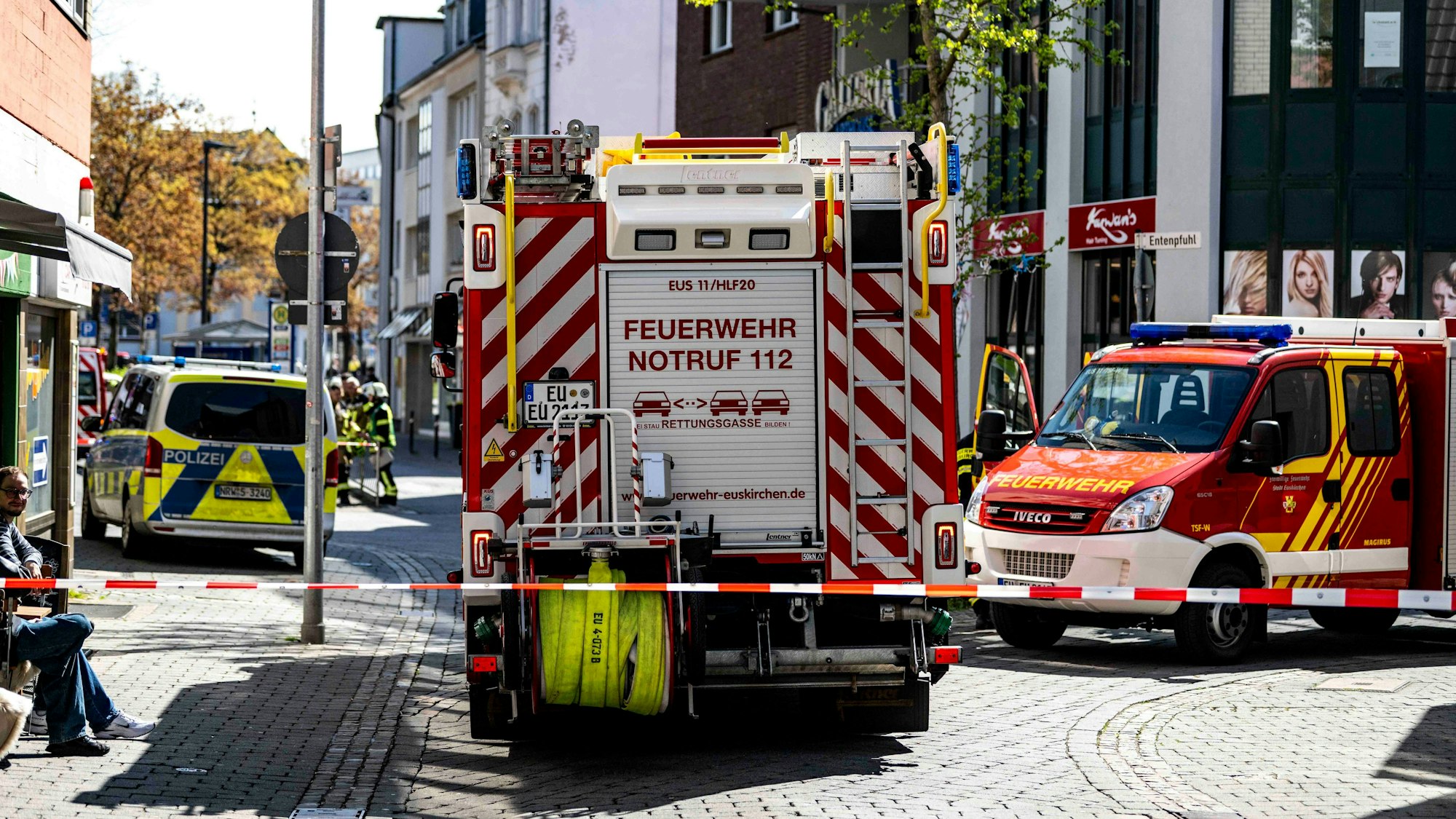 Das Bild zeigt zwei Feuerwehrfahrzeuge und ein Polizeiauto. Sie stehen hinter rot-weißem Flatterband.