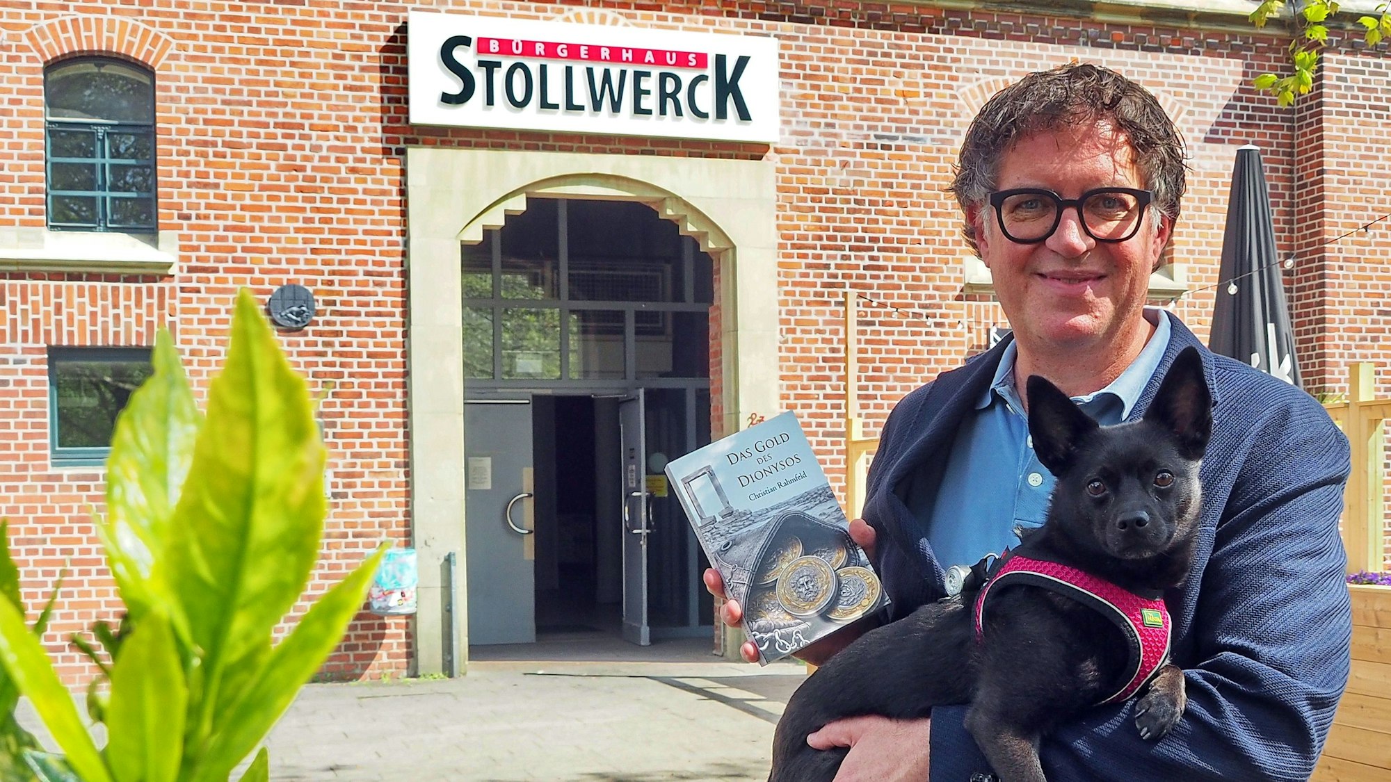 Ein Mann mit schwarzem Haar und Brille steht vor einem Backsteingebäude, ein Buch in der Hand und einen kleinen Hund auf dem Arm.