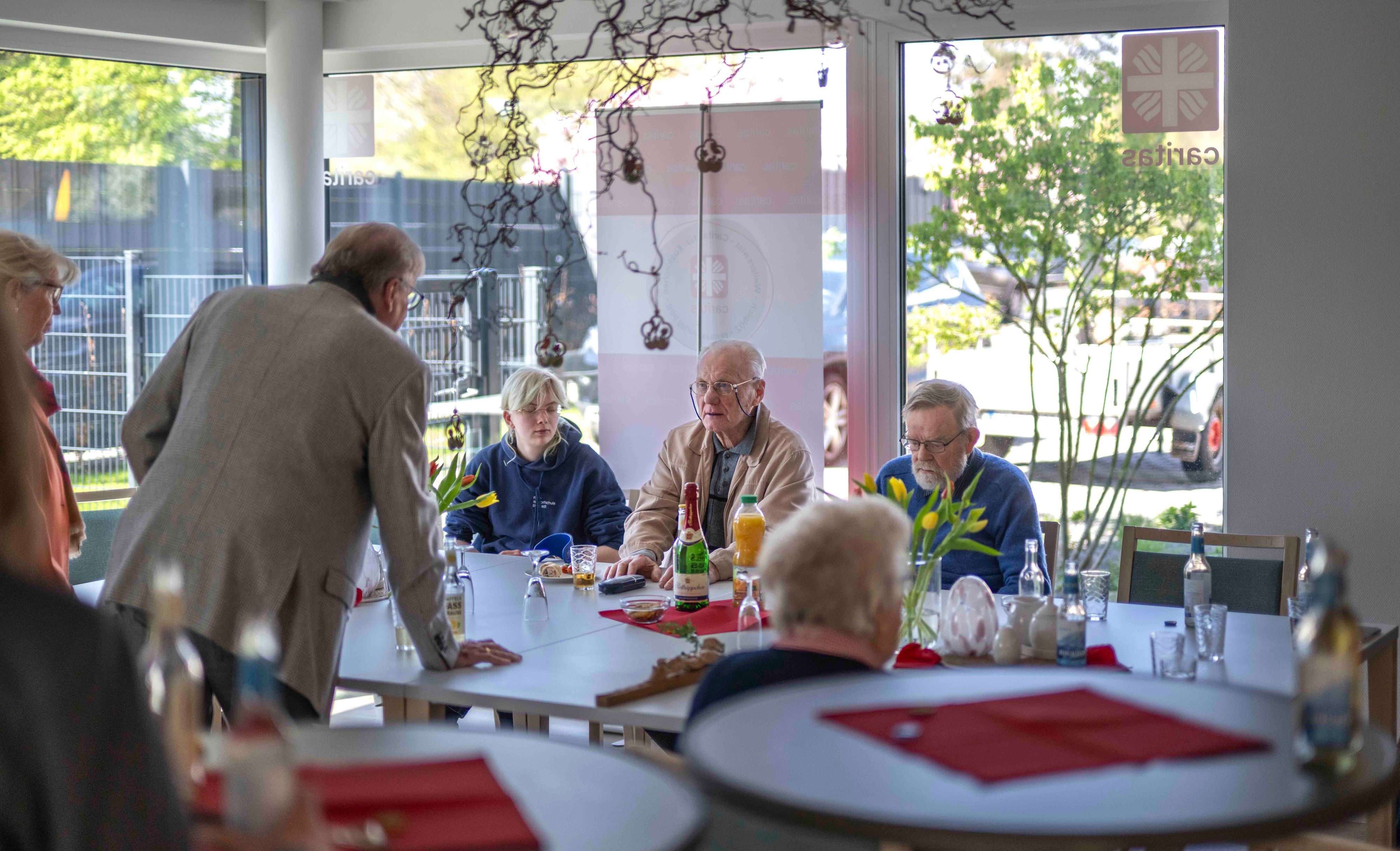 Blick in die lichthelle Tagesstätte. Ein paar Gäste sitzen am Tisch, der mit Blumen geschmückt ist.