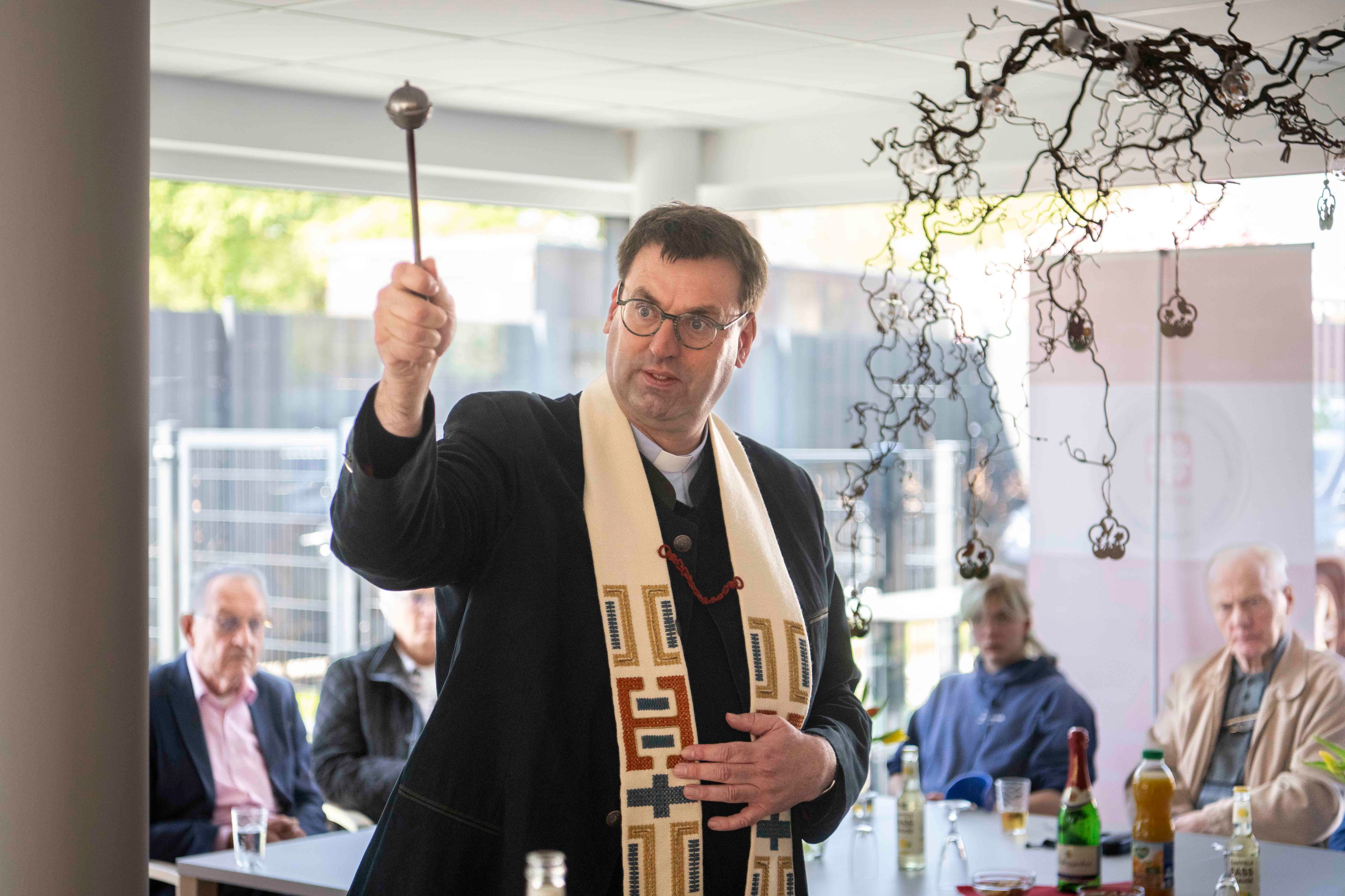 Kreisdechant Guido Zimmermann spritzt mit einem Aspergill Weihwasser in die Tagesstätte, im Hintergrund sitzen Gäste der Feier.