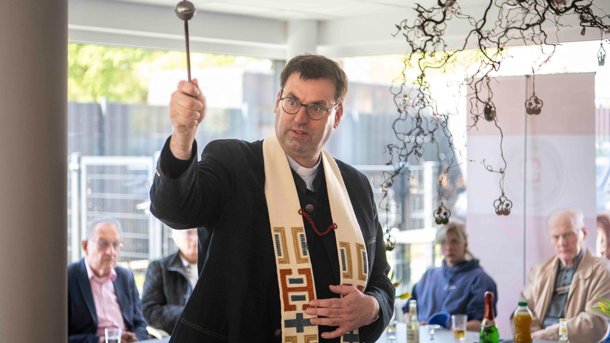 Kreisdechant Guido Zimmermann spritzt mit einem Aspergill Weihwasser in die Tagesstätte, im Hintergrund sitzen Gäste der Feier.