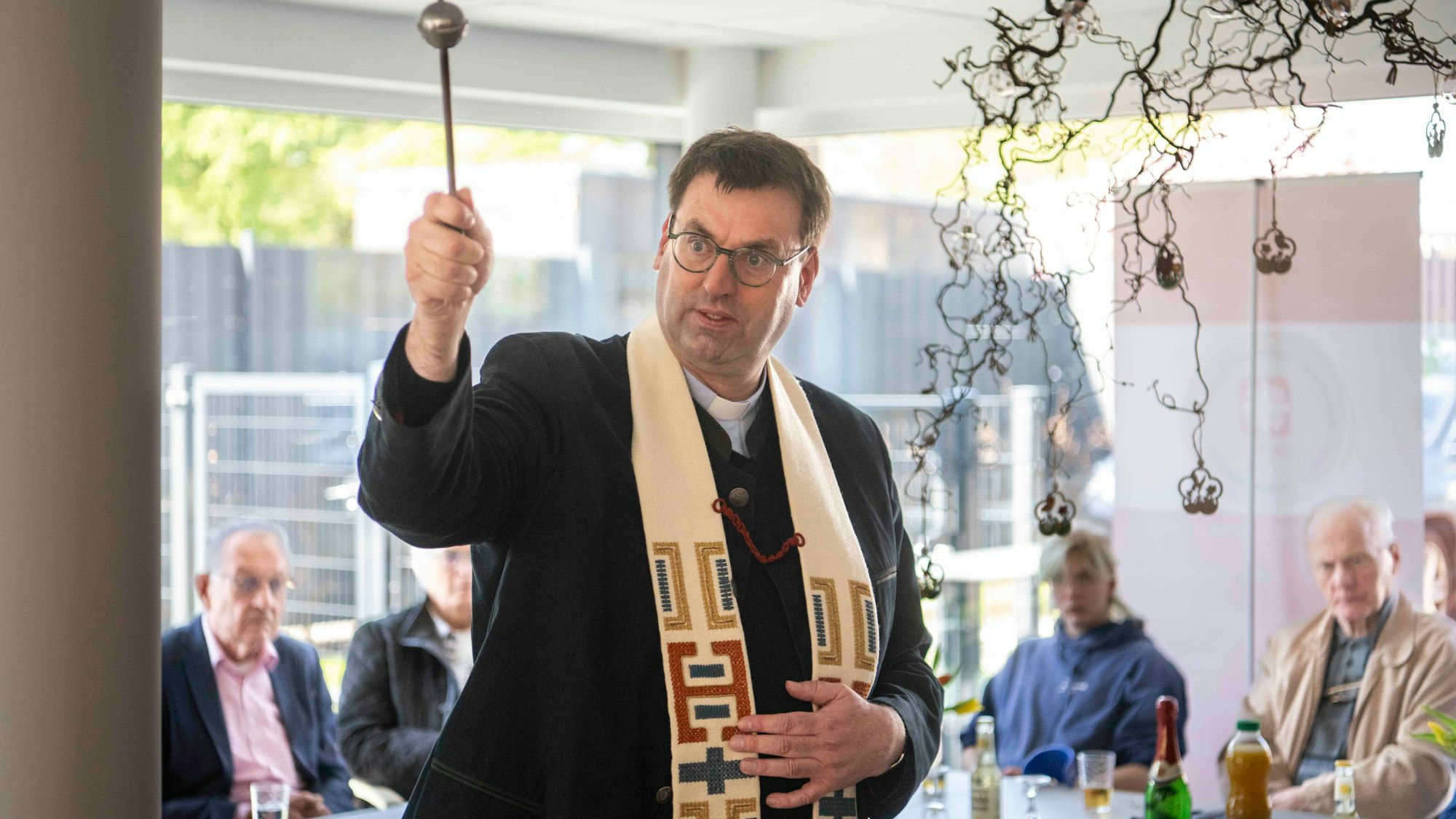 Kreisdechant Guido Zimmermann spritzt mit einem Aspergill Weihwasser in die Tagesstätte, im Hintergrund sitzen Gäste der Feier.