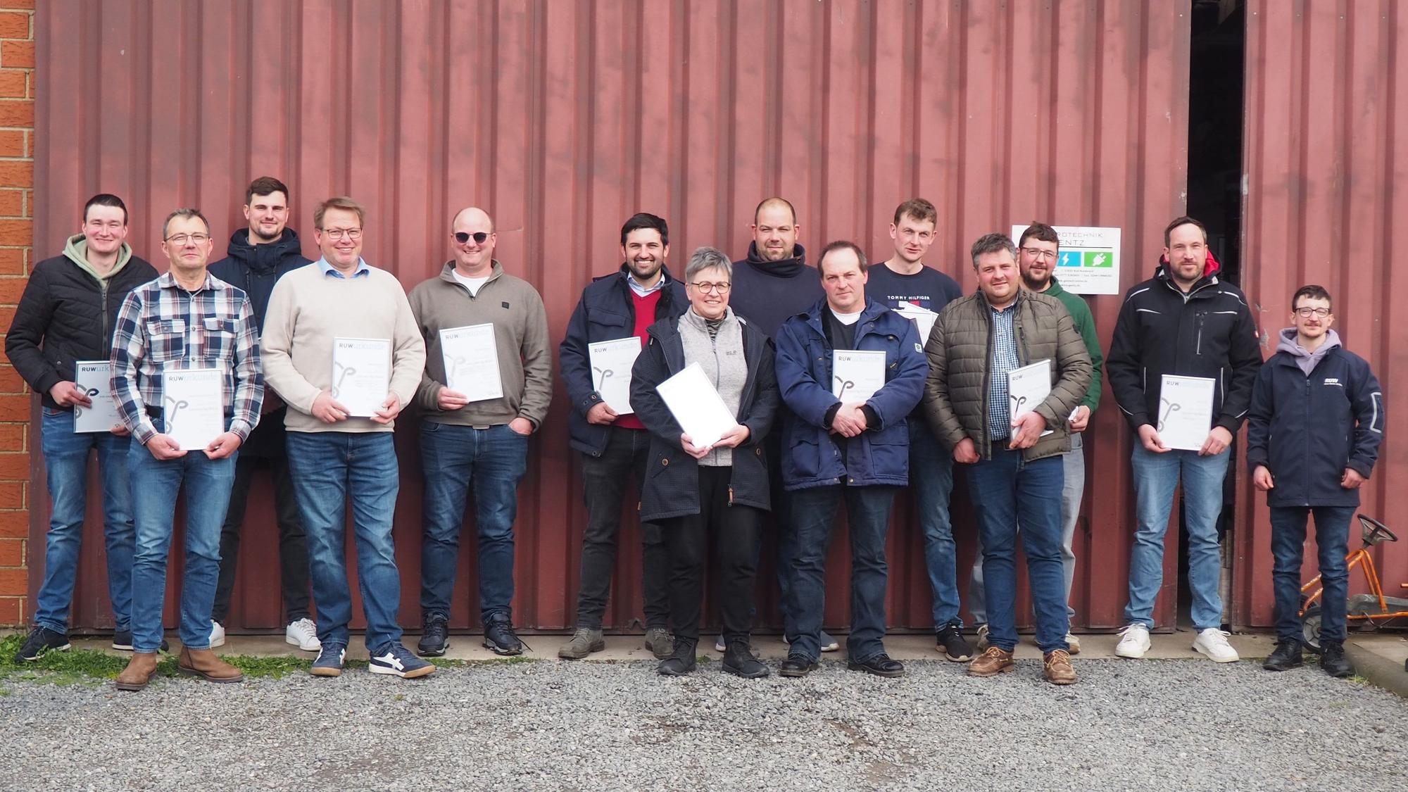 Eine Gruppe von 14 Landwirten, darunter eine Frau, steht mit Urkunden in der Hand vor einem roten Metalltor.
