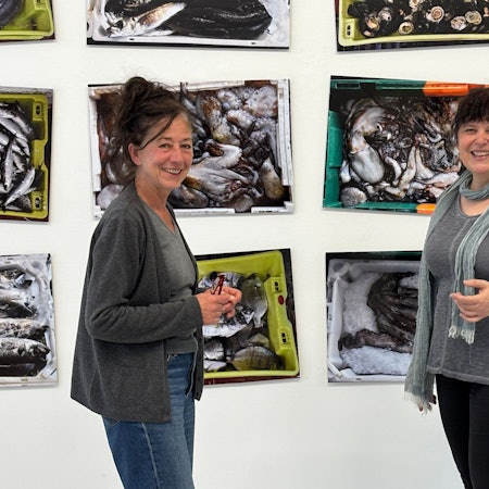 Die Obengenannten stehen lächelnd vor Bildern, die tote Fische und Oktopusse in Kisten zeigen.