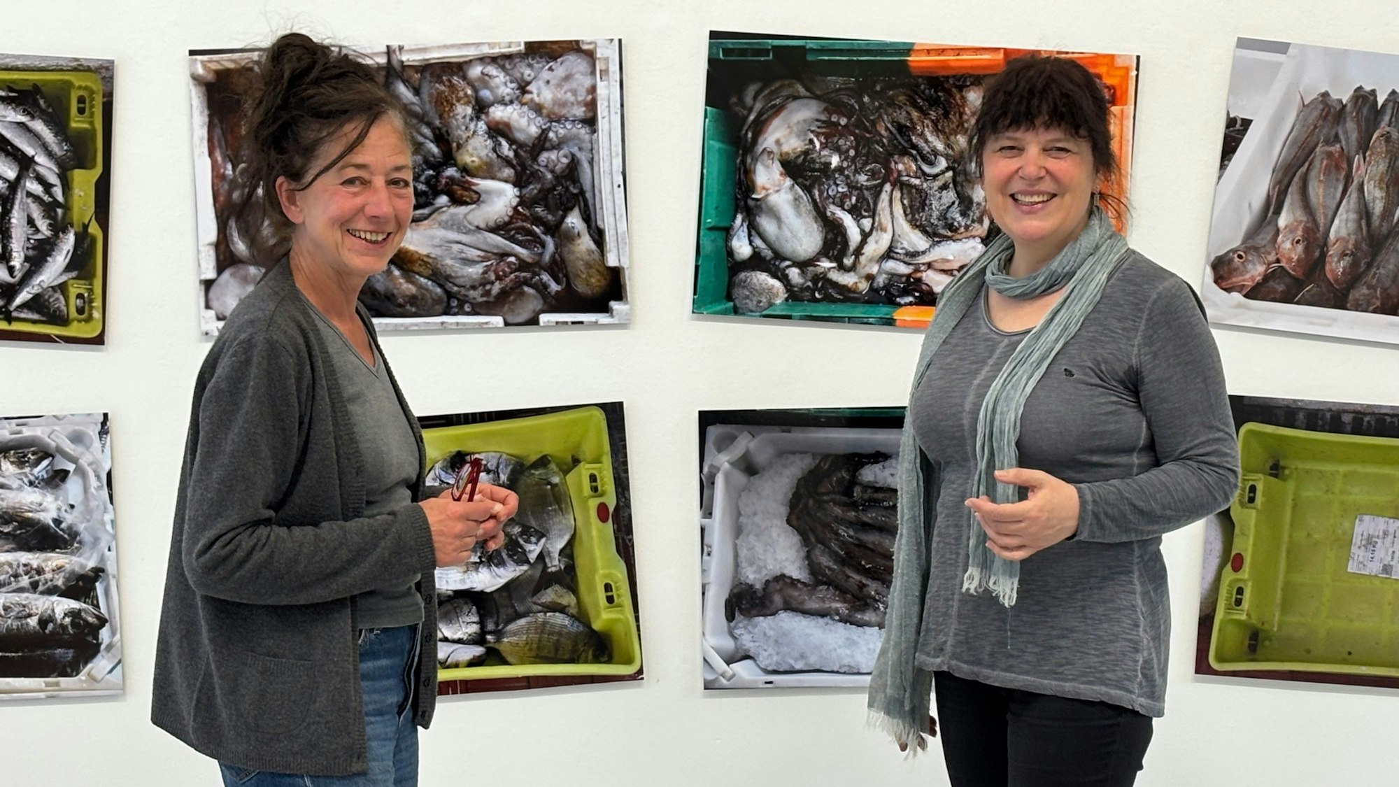 Die Obengenannten stehen lächelnd vor Bildern, die tote Fische und Oktopusse in Kisten zeigen.
