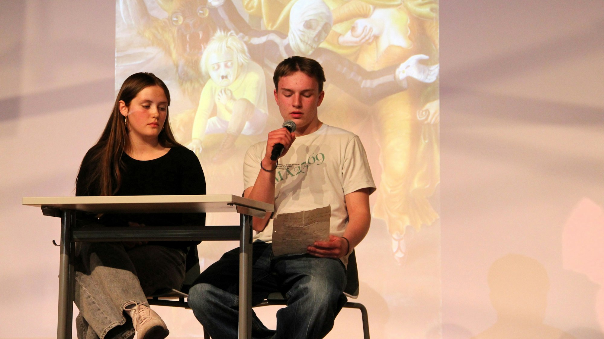 Ein Mädchen und ein Junge sitzen an einem Tisch, der junge Mann liest etwas vor.