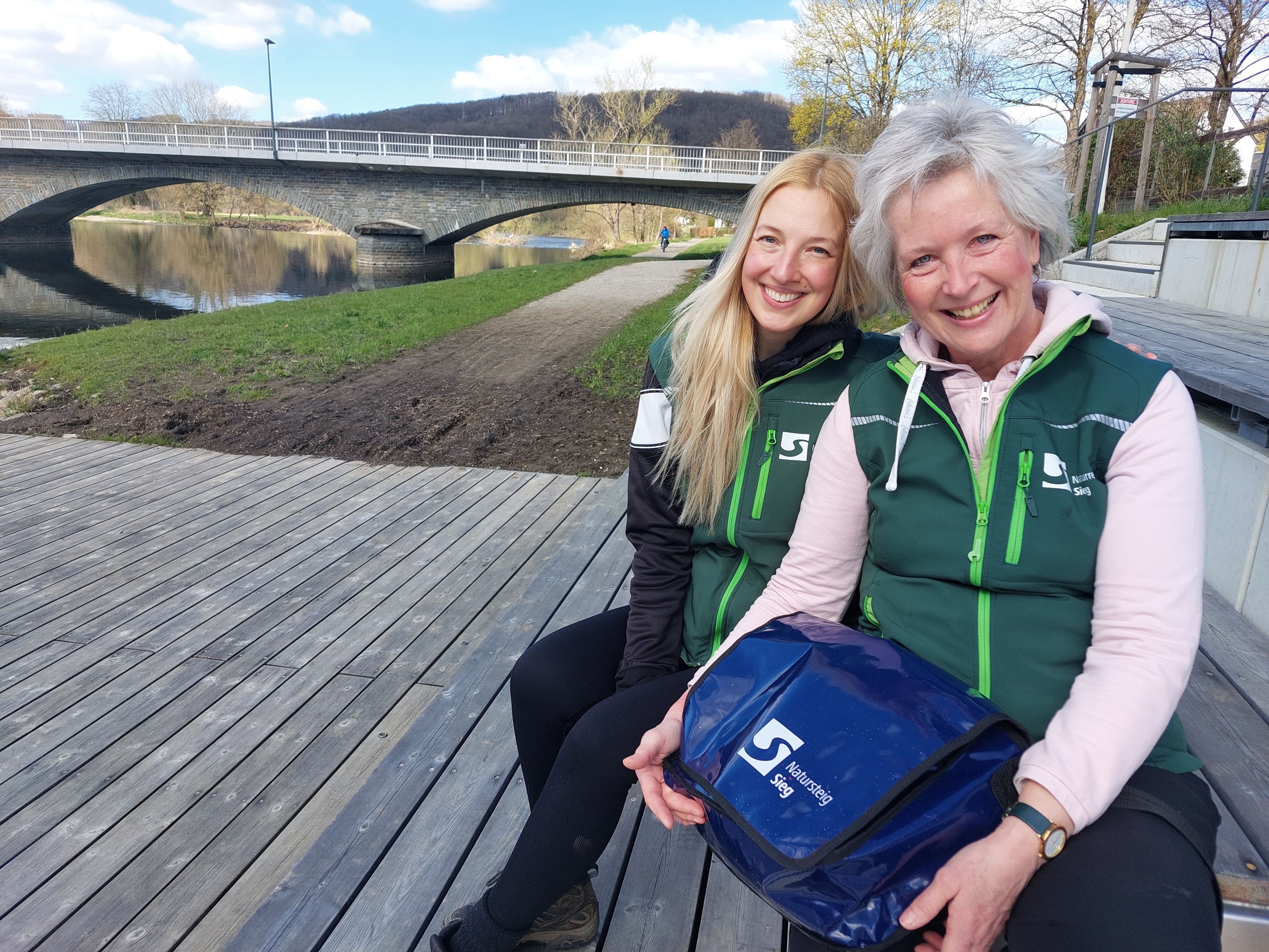 Zwei Frauen sitzen unweit einer Brücke. Sie tragen grüne Westen.
