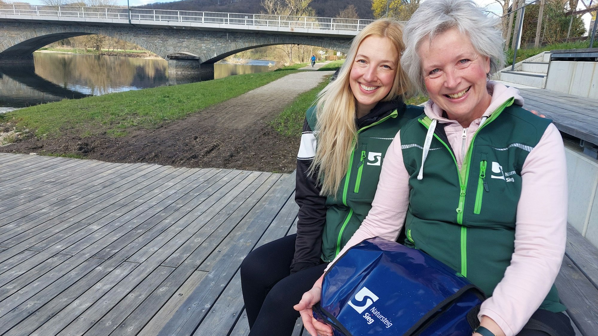 Zwei Frauen sitzen unweit einer Brücke. Sie tragen grüne Westen.