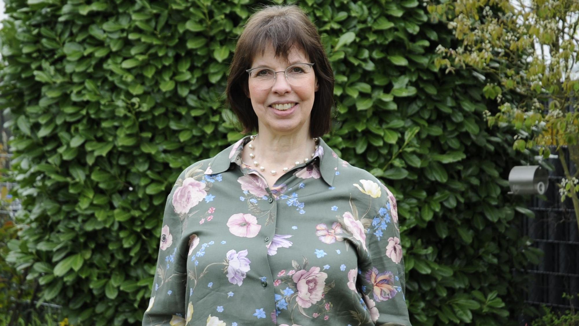 Eine Frau mit dunklen Haaren steht vor einer Hecke.