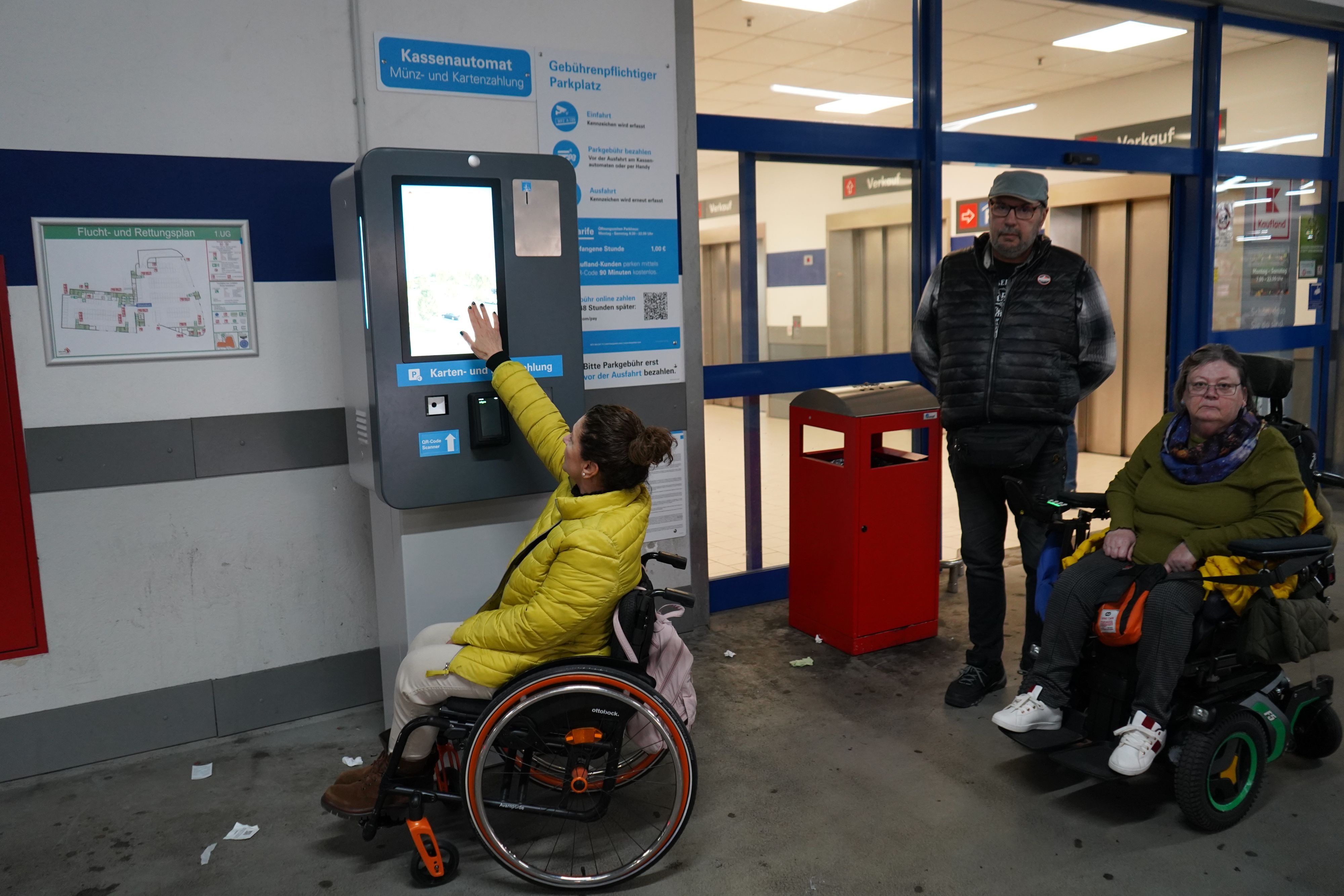 Zu sehen sind zwei Frauen im Rollstuhl sowie ein Mann, sie befinden sich an einem Parkautomaten.