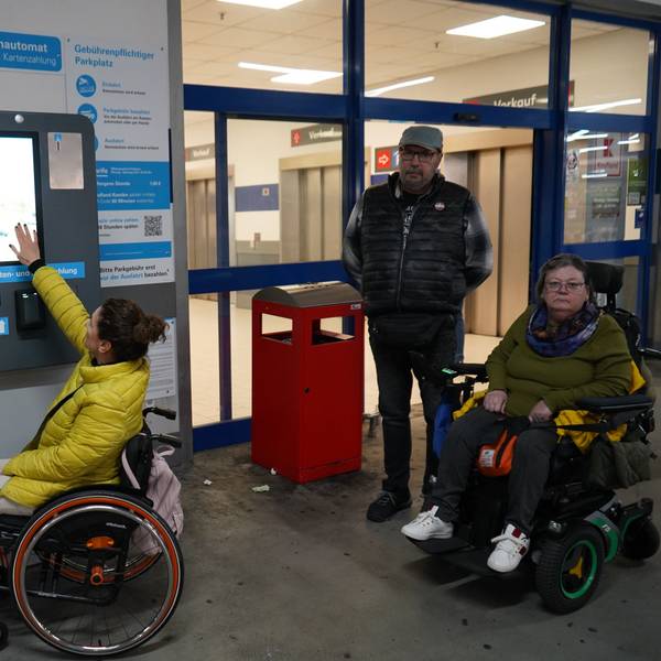 Zu sehen sind zwei Frauen im Rollstuhl sowie ein Mann, sie befinden sich an einem Parkautomaten.