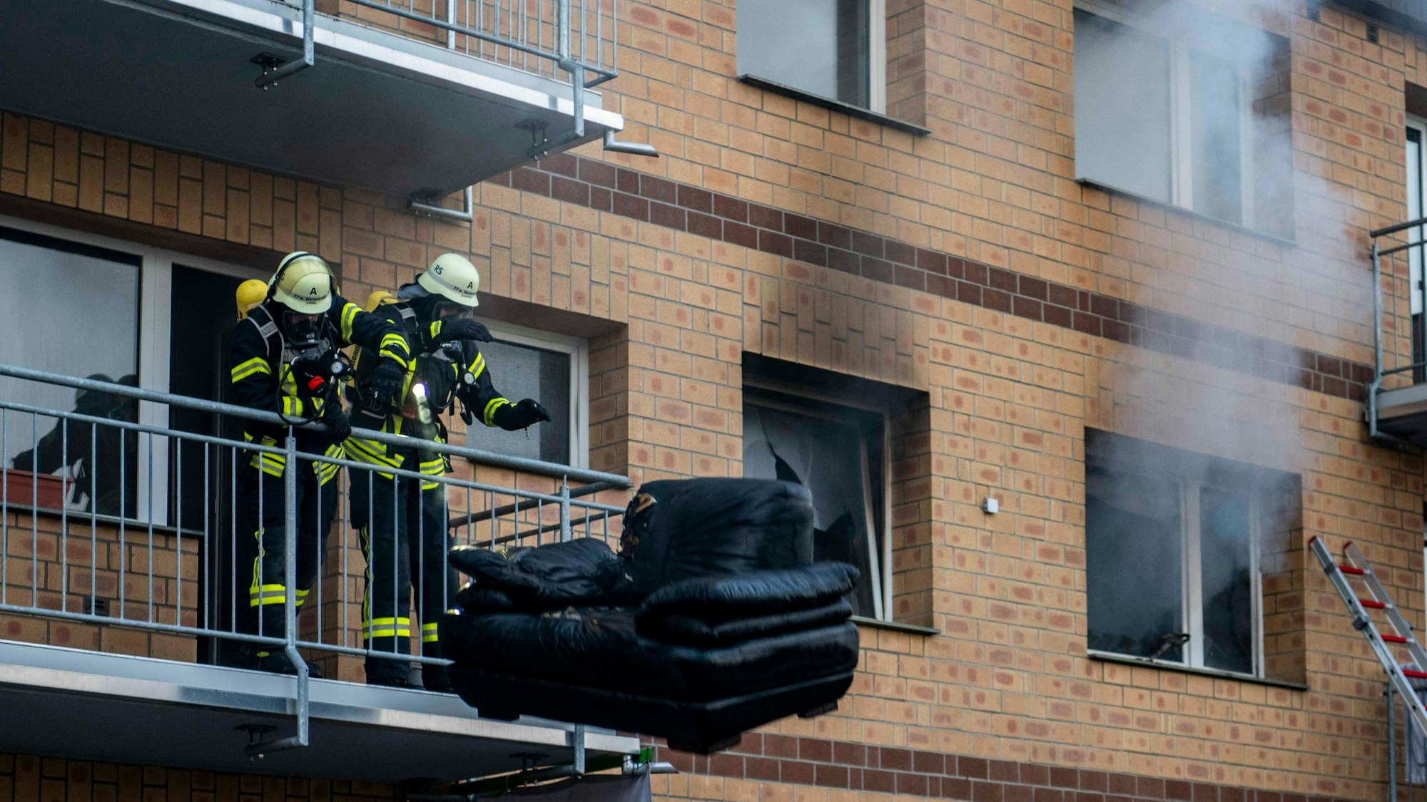 Das Bild zeigt zwei Feuerwehrleute, die einen Sessel vom Balkon werfen, um ihn von Kollegen ablöschen zu lassen.
