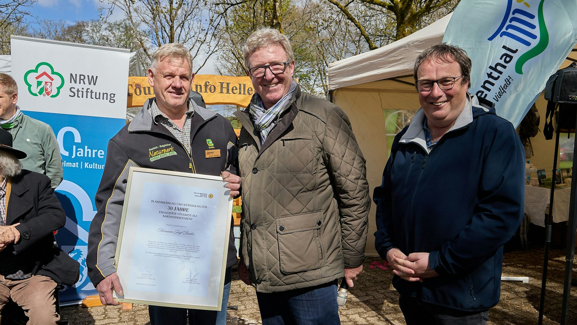 Drei Männer stehen vor verschiedenen Aufstellern nebeneinander, einer präsentiert eine Urkunde.