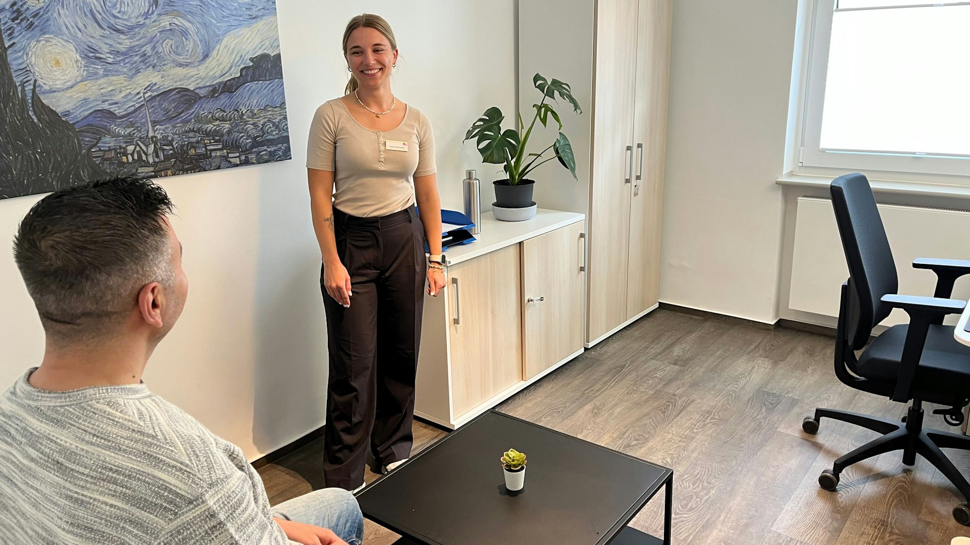 In einem Beratungszimmer sitzt ein junger Mann an einem Tisch, eine junge Frau steht im Raum.
