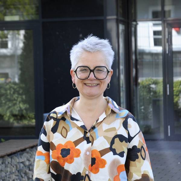 Eine Frau mit Brille und kurzen Haaren steht vor einem Gebäude.