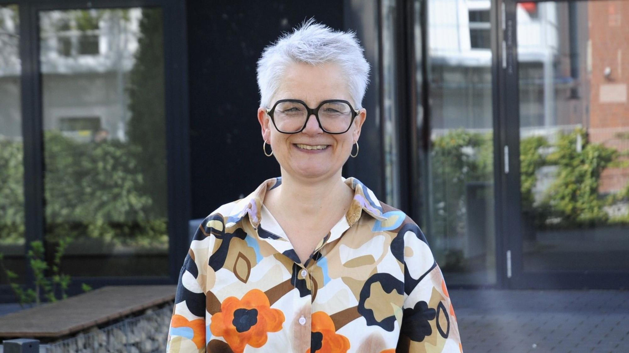 Eine Frau mit Brille und kurzen Haaren steht vor einem Gebäude.