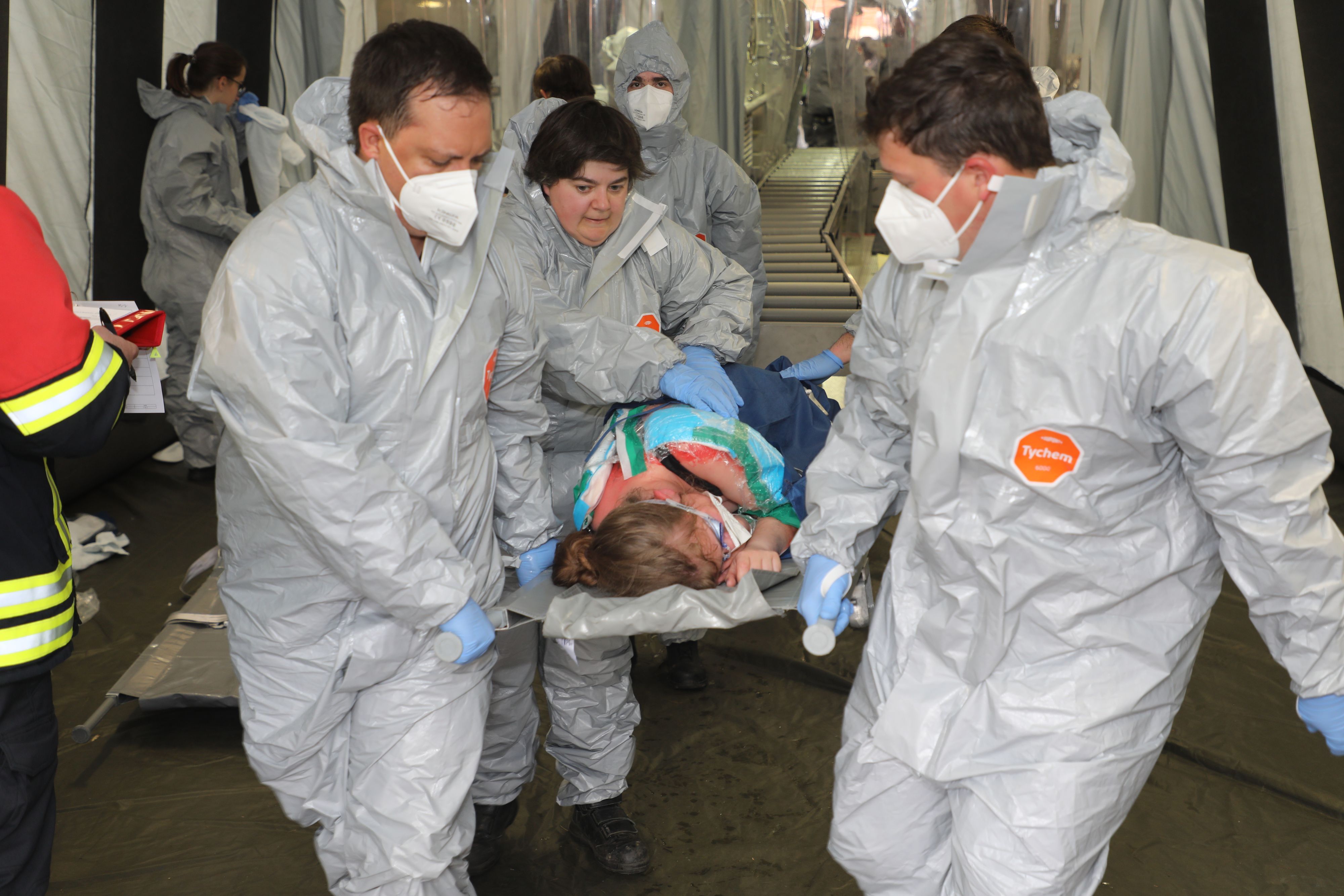 Vier Einsatzkräfte in Schutzkleidung trage eine Verletzte mit Verätzungswundern auf einer Trage aus der Dekontamination.