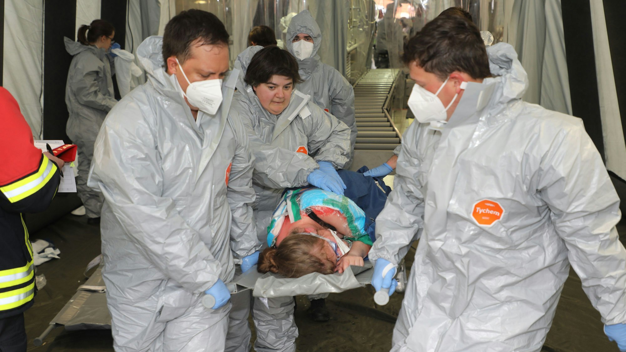 Vier Einsatzkräfte in Schutzkleidung trage eine Verletzte mit Verätzungswundern auf einer Trage aus der Dekontamination.