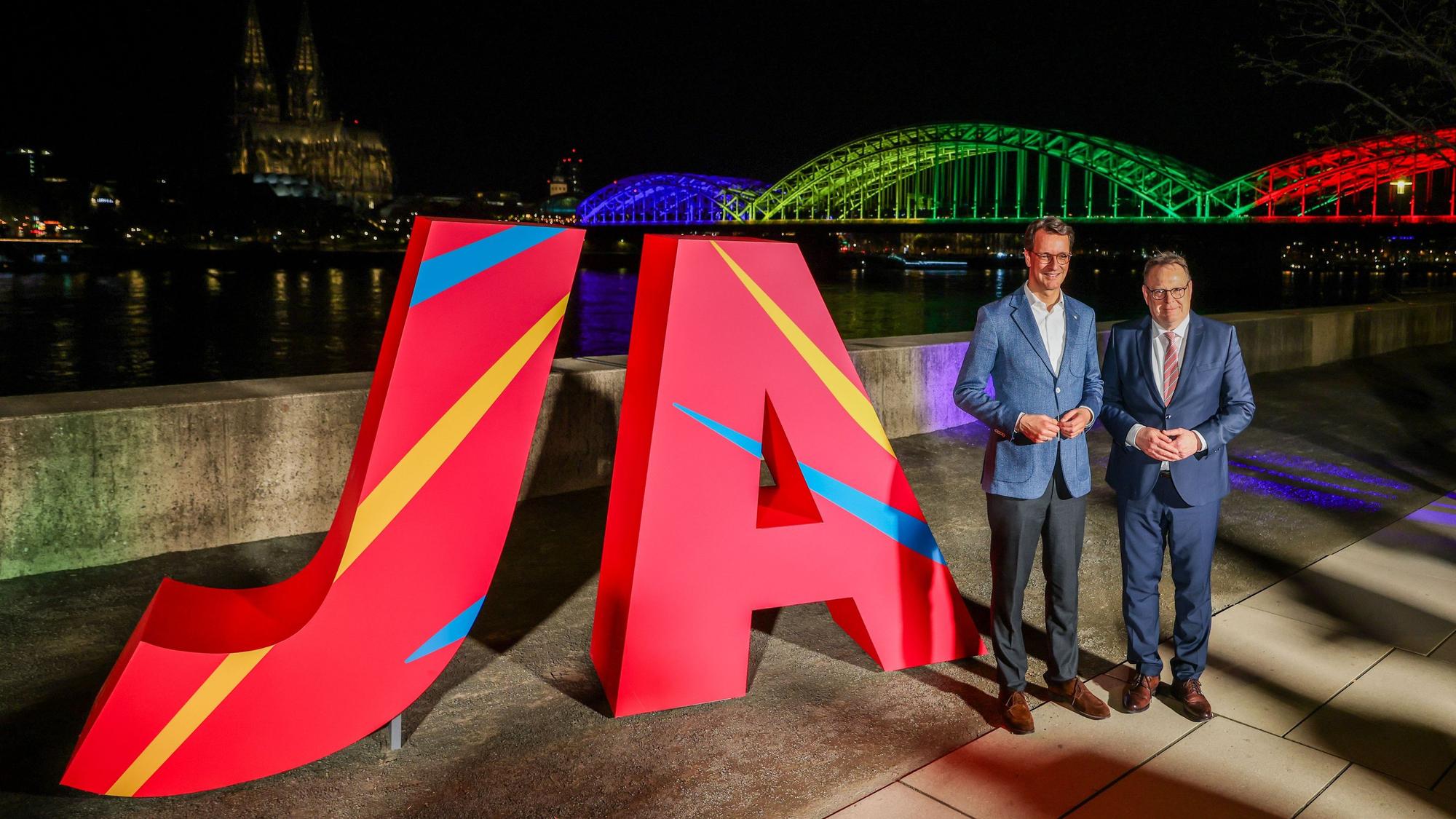 Hendrik Wüst (CDU, l.), NRW-Ministerpräsident, und Torsten Burmester (SPD), Oberbürgermeister der Stadt Köln, stehen vor dem großen „Ja“ und der in den olympischen Farben angeleuchteten Hohenzollernbrücke.
