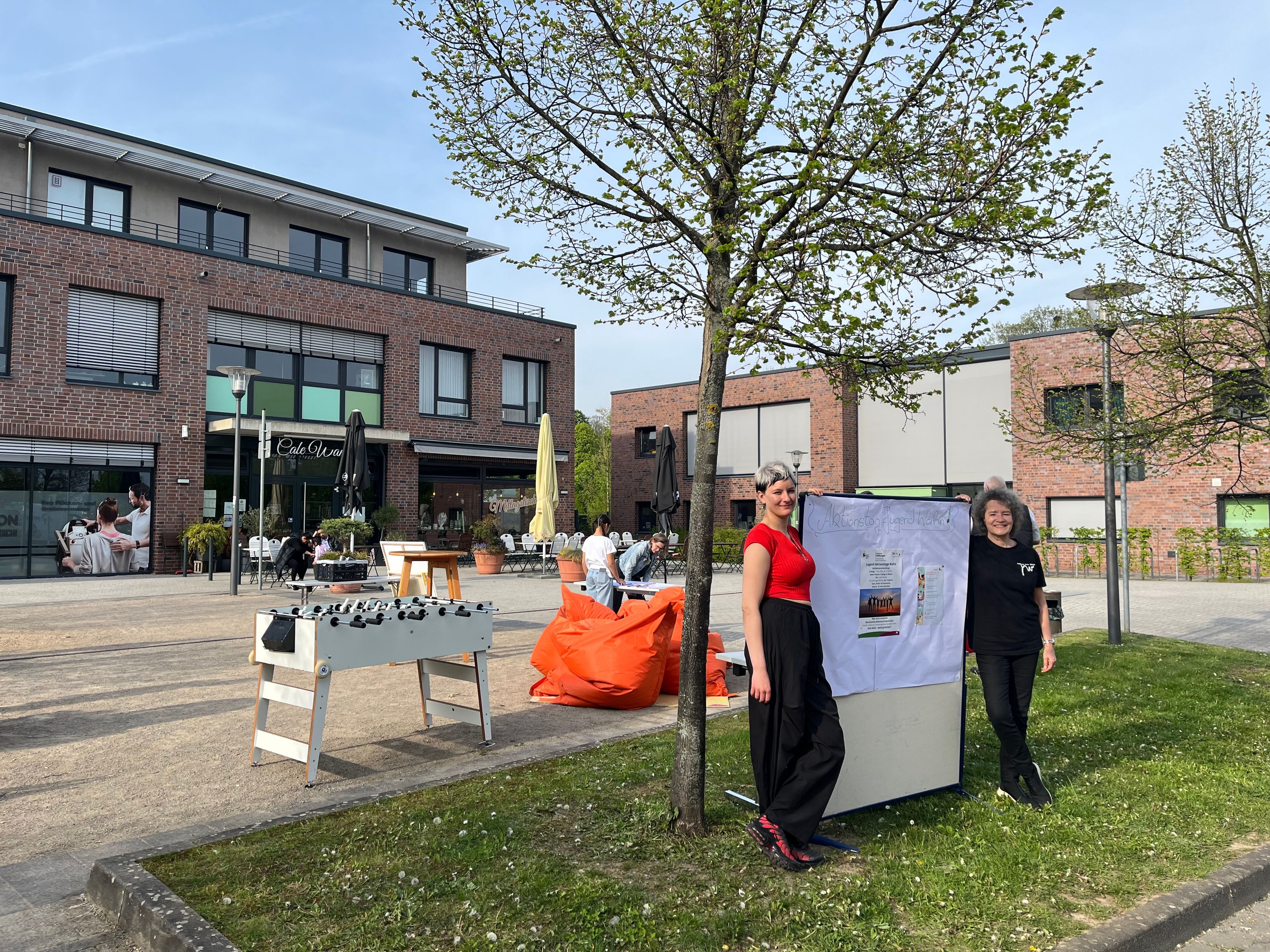 Zwei Frauen stehen links und rechts von einem Aufsteller. Dahinter ist ein großer Platz zu sehen, auf dem ein Kickertisch steht und Stühle und Tische.