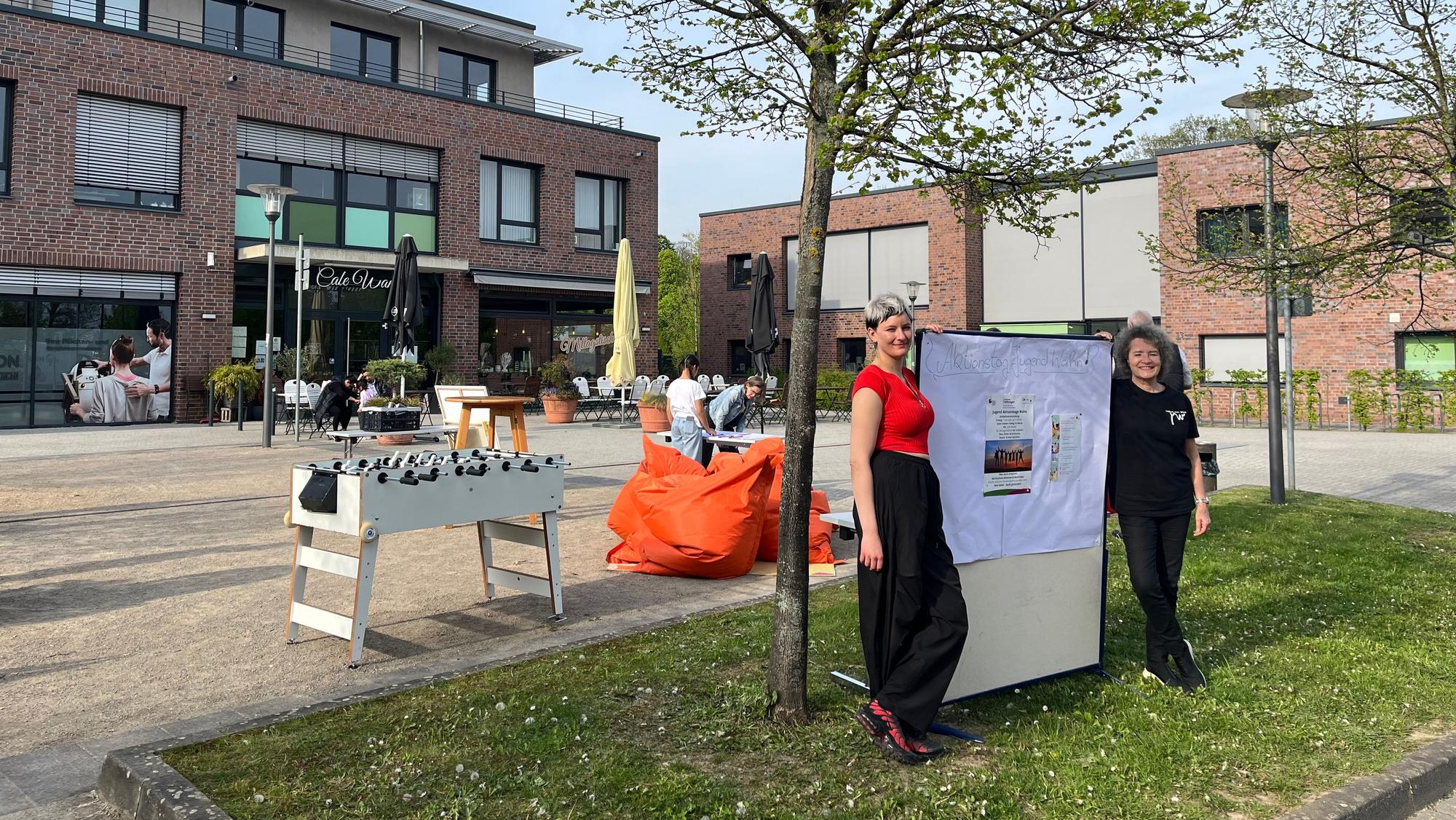 Zwei Frauen stehen links und rechts von einem Aufsteller. Dahinter ist ein großer Platz zu sehen, auf dem ein Kickertisch steht und Stühle und Tische.