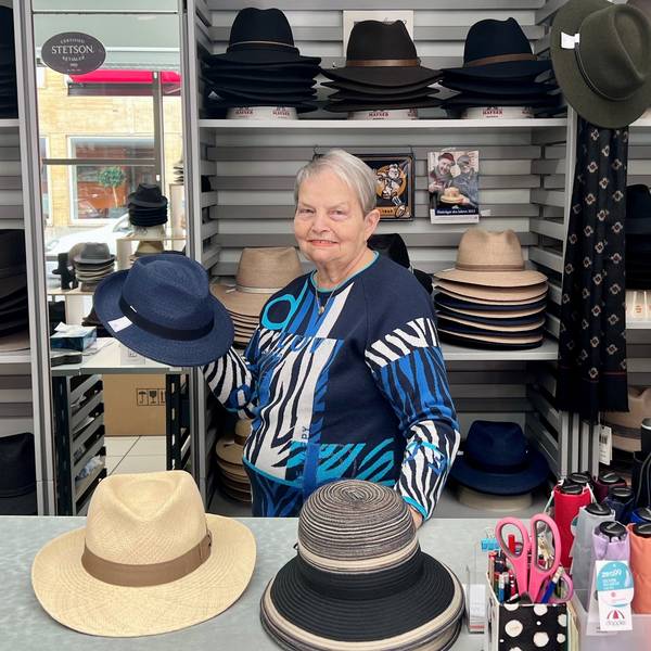 Ein ältere Dame steht hinter einem Verkaufstresen, sie hält einen blauen Herrenhut in der Hand.