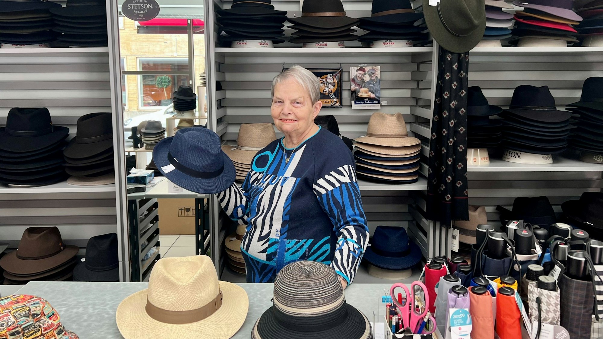 Ein ältere Dame steht hinter einem Verkaufstresen, sie hält einen blauen Herrenhut in der Hand.