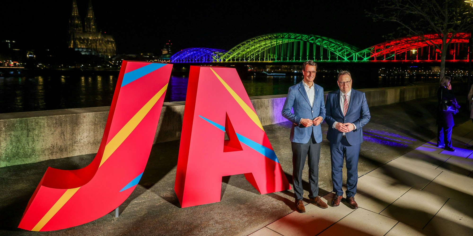 Ministerpräsident Hendrik Wüst (l.) und Kölns OB Torsten Burmester vor der beleuchteten Hohenzollernbrücke.